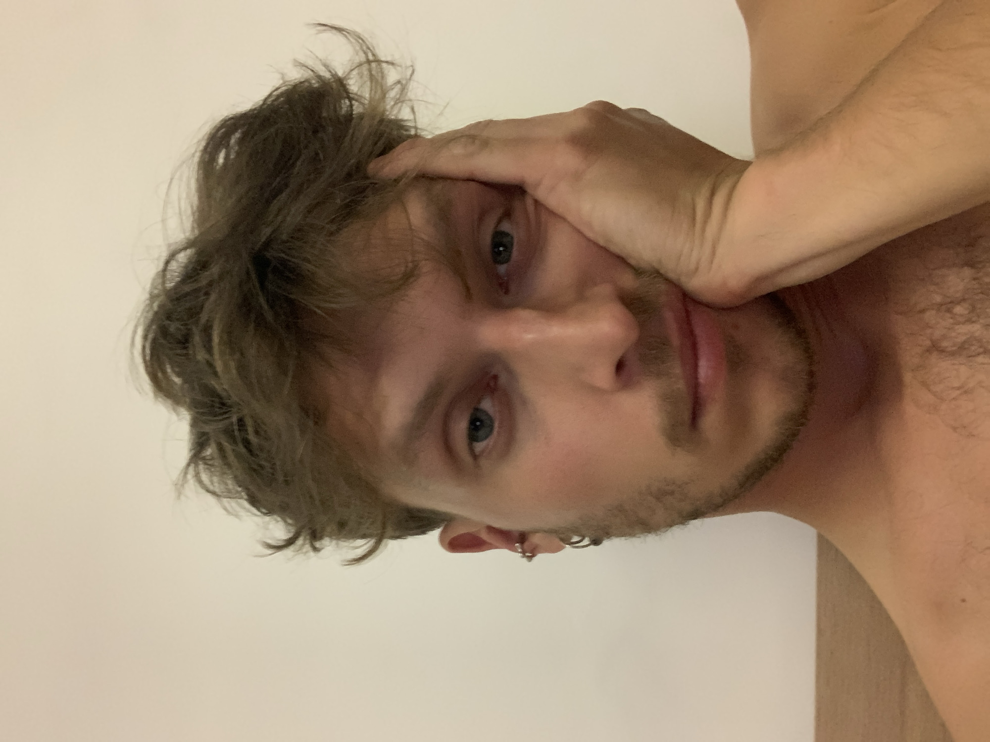 A close-up portrait shows a man, appearing to be in Tel-Aviv, Israel, against a plain, light-colored wall. The man has messy, light brown hair, blue eyes, and light stubble on his face. He is looking directly at the camera with a neutral, slightly pensive expression. His right hand is resting casually on the side of his head, covering part of his forehead. He has two small hoop earrings visible in his left ear. The man is bare-chested, revealing some body hair on his chest and shoulder. The lighting is soft and even, typical of indoor artificial light. No other objects or text are visible in the frame.