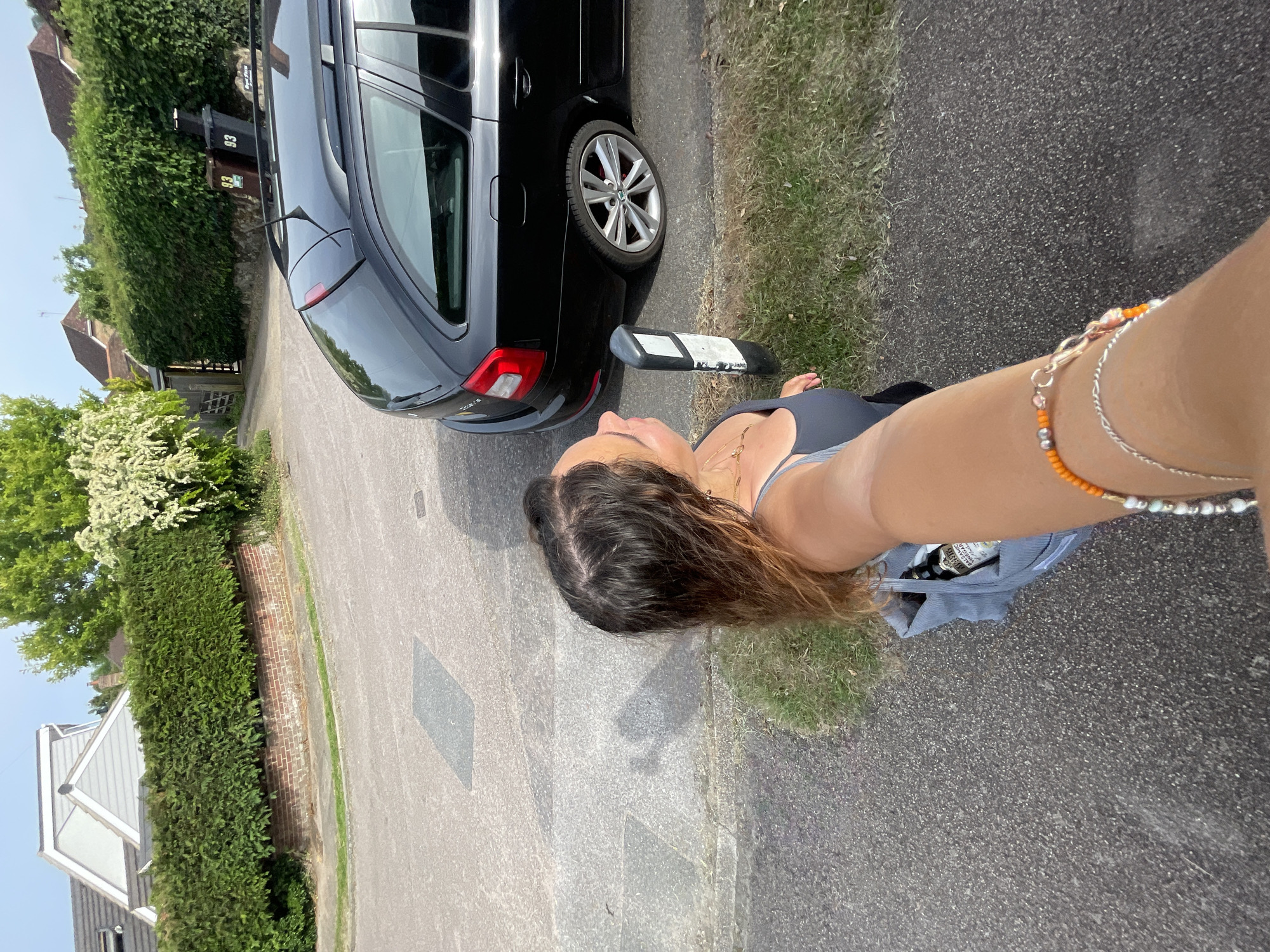 A sideways outdoor shot shows a woman standing on a paved road next to a black car and a grassy verge. The woman is wearing a grey top and denim shorts. She holds up her arm, adorned with bracelets, and seems to be taking a selfie. Behind her is the rear of a black Skoda estate car parked on the side of the road. There is a small black and white bollard or marker next to the car. The location appears to be a residential street with hedges and houses visible in the background. Based on the visible garbage cans marked with “93” and "Maidstone Borough Council", the location is possibly within the city of Maidstone, United Kingdom.