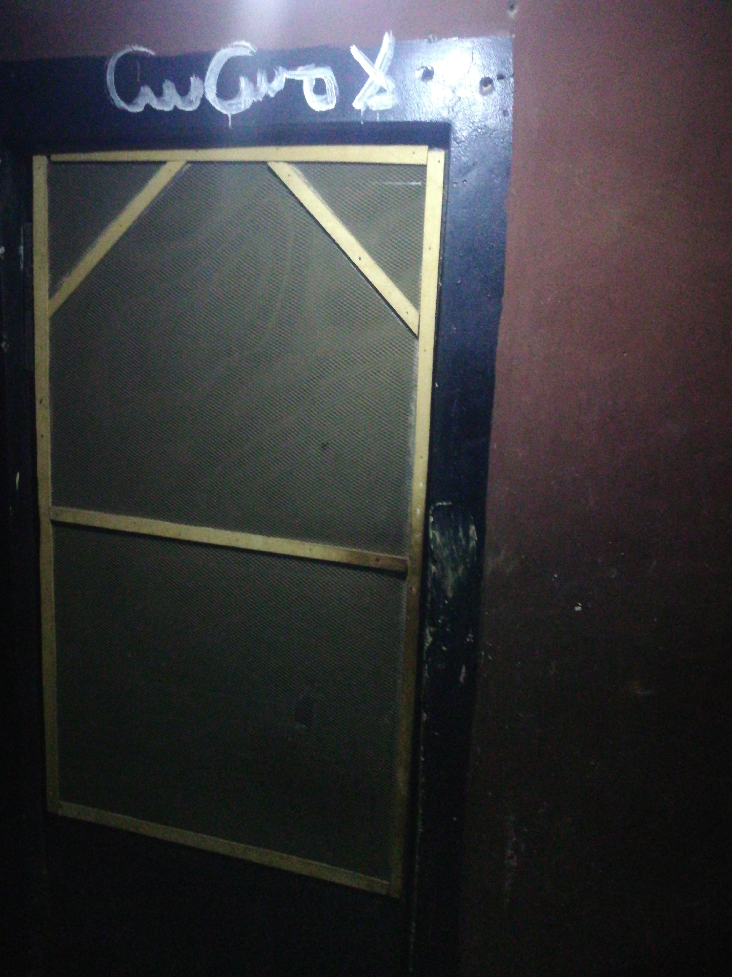 The image depicts a dark and seemingly neglected interior space. A rectangular window or screen is prominent, featuring a wooden frame and a dark, woven mesh. Support beams reinforce the top portion of the frame, forming a triangular shape. Above the window, white graffiti or writing is visible on the dark wall. The surrounding wall is painted in two tones: a dark, possibly black, color around the window and a dark maroon or brown on the right side. The overall lighting is dim, contributing to the somber and somewhat dilapidated atmosphere of the scene. The location in the city is Unknown, Unknown.