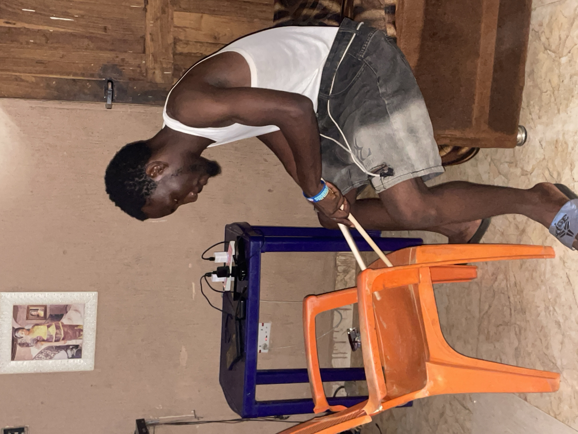 A man with dark skin and a beard, wearing a white tank top and grey shorts, is sitting on a brown sofa. He is holding two light-colored drumsticks in his hands, with the tips resting on an orange plastic chair. In the background, a blue table holds several electronic devices and chargers. A framed picture of two people hangs on a beige wall. The scene appears to be indoors, possibly in a residential setting in Lokoja, Nigeria. The lighting is dim, suggesting it may be evening or nighttime.