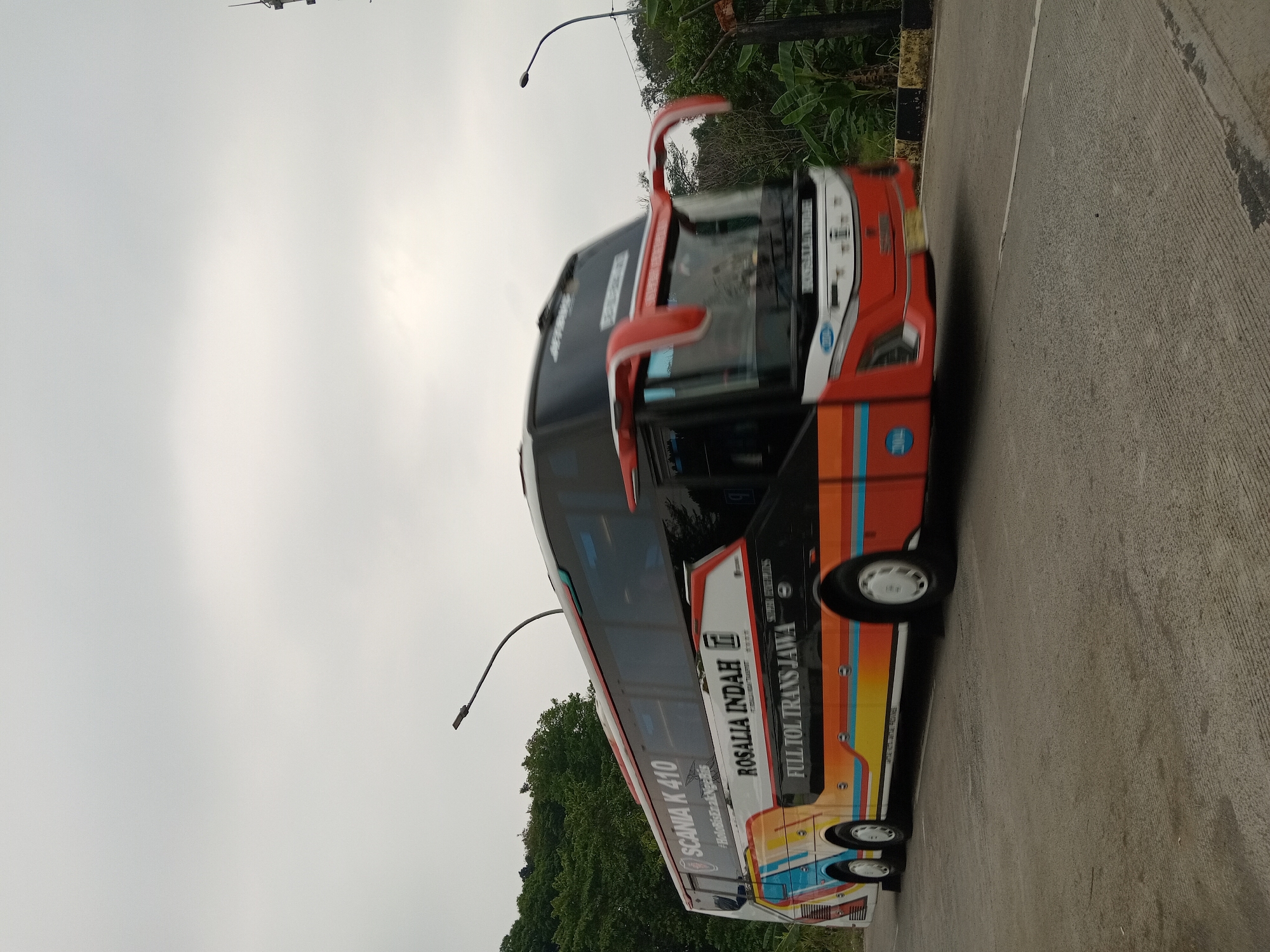 An external, eye-level, full-shot photograph captures a double-decker bus in motion on a paved road under an overcast sky. The bus, predominantly orange with a black top deck, is adorned with colorful stripes of yellow, blue, and red along its side. Text on the bus reads "SCANIA K 410," "ROSALIA INDAH," and "FULL TOL TRANS JAWA." The bus is positioned on the right side of the frame, moving from left to right. Lush green trees and foliage line the left side of the road, and a blurred fence and banana trees are visible on the right. The road surface is a light gray concrete with visible expansion joints. Two streetlights are present in the frame: one a modern pole with a curved arm extending over the road on the left, and another on the right with a similar curved arm. The image appears to have been taken in Depok, Indonesia, based on the visible branding and context.