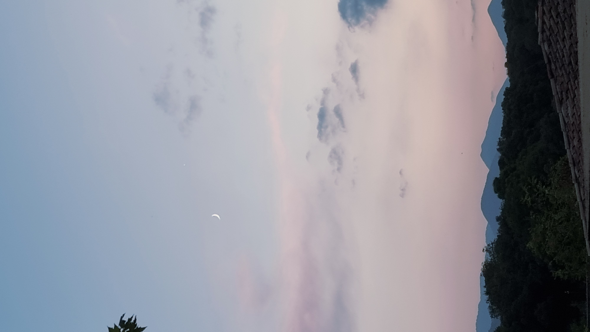This image depicts a twilight sky over a mountainous landscape. The sky transitions from a pale blue on the left to soft pink and lavender hues as it stretches towards the right, suggesting either sunrise or sunset. Scattered clouds, some appearing wispy and others darker and more defined, drift across the sky. A crescent moon is visible in the upper left portion of the frame.
In the foreground, the silhouette of dark green trees and foliage forms a dense band along the base of the mountains. Beyond the trees, a series of layered mountains recede into the distance, their outlines softened by the atmospheric haze. The distant mountains appear in shades of blue, indicating their depth and distance. On the far right edge of the image, a section of a tiled roof suggests a man-made structure, possibly a building or house, overlooking the natural scenery. The overall mood is serene and tranquil, characteristic of the quiet moments between day and night. The image appears to be taken from a slightly elevated viewpoint. No text is visible in the image.