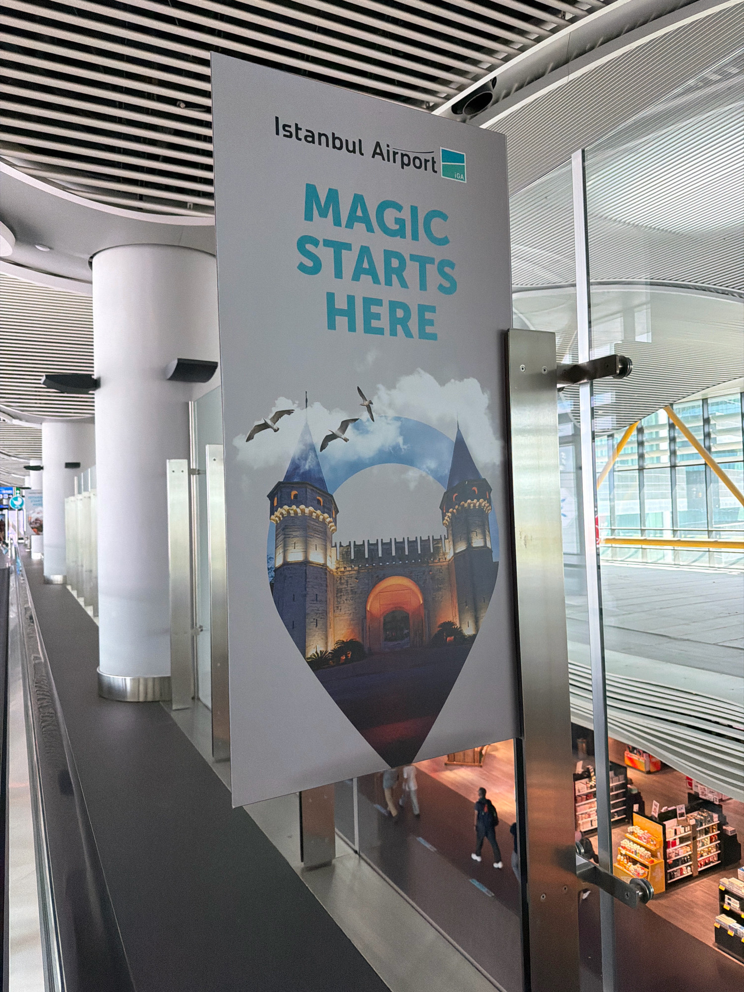 The image shows a large sign hanging in a modern airport terminal. The sign features the text "Istanbul Airport" at the top, followed by "MAGIC STARTS HERE" in large, teal-colored letters. Below the text is an image of a historical Turkish castle with two turrets and a fortified wall, illuminated by warm lights, with three seagulls flying in a cloudy sky above it. The sign is positioned in front of a glass wall, revealing a multi-level interior with shops and people visible below. The architectural style of the airport is characterized by clean lines, white pillars, and a slatted ceiling. The airport is located in Arnavutköy, Turkey.
