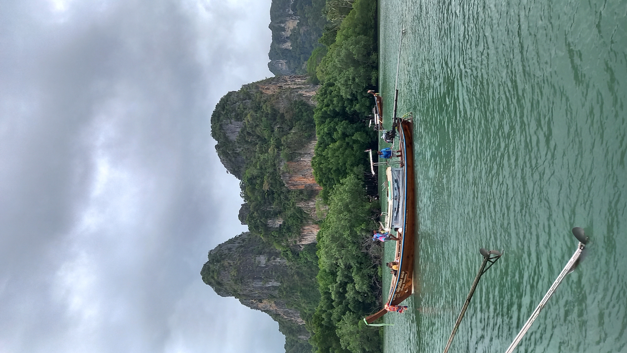 The image shows a longtail boat on the water in Ban Ao Nang, Thailand. The boat has several people on board. In the background are limestone cliffs covered in dense vegetation. The sky is overcast. The water is green. Some long metal poles are sticking out of the water in the foreground.