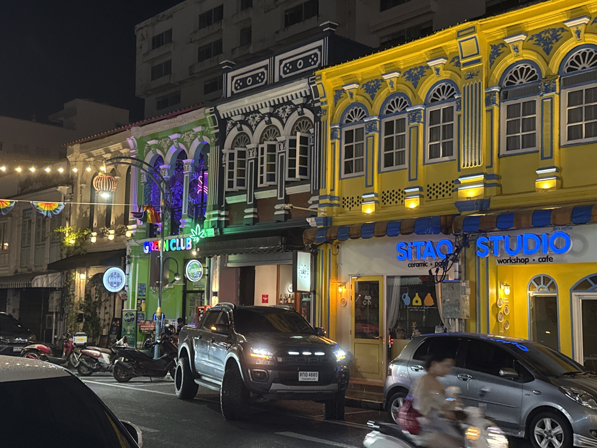 This is a night-time street view in Phuket City Municipality, Thailand, featuring a row of colorful buildings with ornate architecture. The buildings are painted in vibrant colors such as green, blue, and yellow, with decorative moldings around the windows and rooftops. Several shops are visible, including one with a neon sign that reads "GREEN CLUB" and another with a blue sign that says "SITAQ STUDIO workshop · cafe". A dark gray pickup truck with its headlights on is parked in the foreground, and several scooters are parked along the sidewalk. A silver hatchback car and a person on a scooter are also visible. The street is illuminated by streetlights and the lights from the shops.