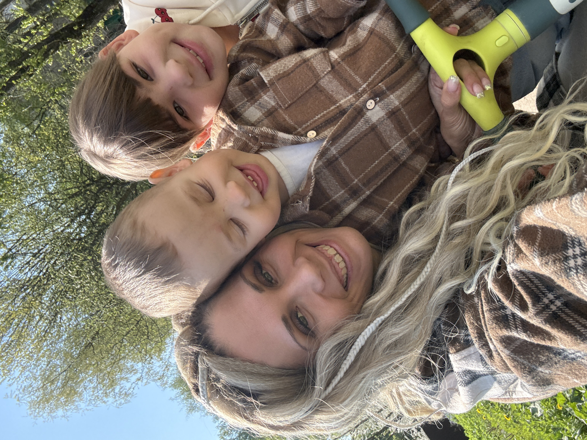 Three people are in the image. A woman with blonde hair and two young children are in the foreground, smiling. The woman is wearing a brown plaid shirt. One child is wearing a brown plaid jacket and a white shirt. The second child is wearing a white sweatshirt. A green and gray object is visible in the upper right. The background includes trees and blue sky. The location is outdoors. It is not possible to identify the specific location as the city of Rusanovo, Russia.