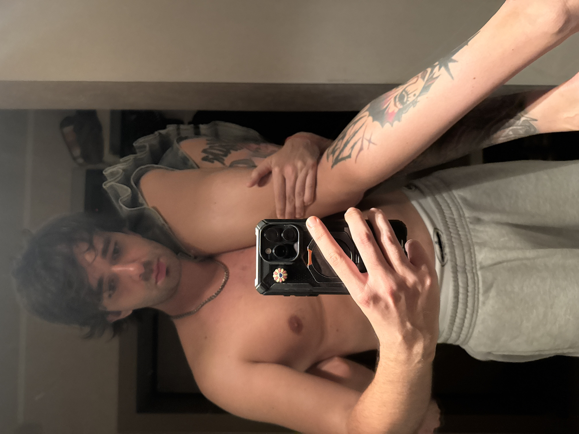 A shirtless person with dark hair and a chain necklace holds a smartphone in their right hand, taking a mirror selfie. They are wearing grey sweatpants. A second person's arm, with visible tattoos, is being held by the first person. The background appears to be an indoor space, possibly a closet or bedroom, with shelves visible. There is no discernible indication of the location being Moscow, Russia.