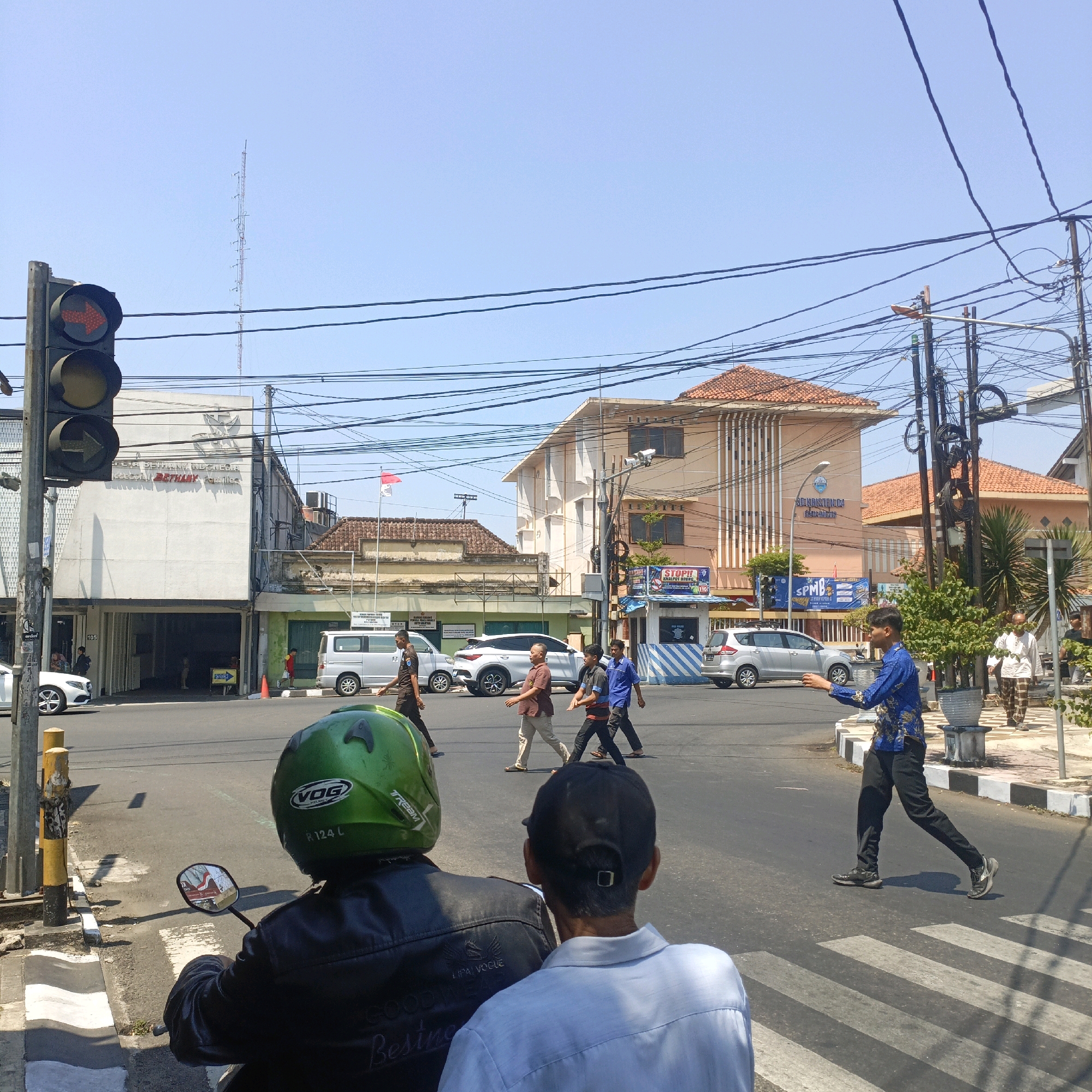 The image depicts a street scene in Salatiga, Indonesia, under a clear blue sky. A traffic light on the left shows a red arrow indicating no right turn. In the foreground, two individuals are seated on a motorcycle, viewed from behind. The rider wears a green helmet, and the person behind wears a light blue shirt and a dark baseball cap.
Across the intersection, several people are walking. Three men in casual attire are crossing the street from right to left, and a man in a blue patterned shirt is walking across the zebra crossing from left to right. To the far right, a man in a white shirt and patterned pants walks away from the camera.
Several vehicles are visible: a white car is parked on the left, and a silver minivan and a white SUV are on the road. The buildings in the background include a multi-story light-colored building with "BETHEL" written on it, and another building with a tiled roof. Power lines crisscross the sky above the street. A flag is visible on a pole attached to one of the buildings. The environment appears to be a commercial or urban area with pedestrian activity.