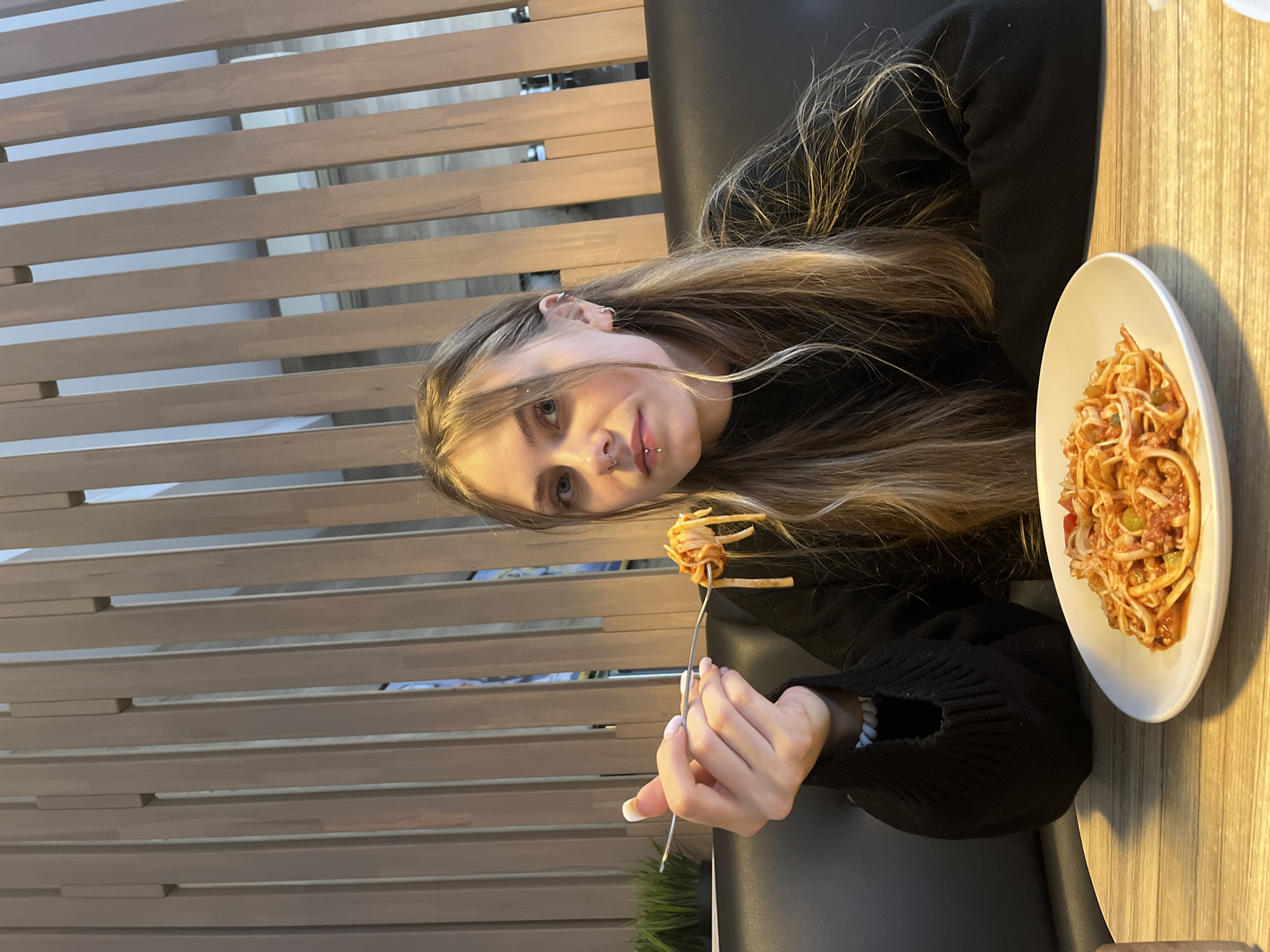 A young female individual is seated at a light-colored wooden table within an indoor commercial establishment in the city of Chaikovsky, Russia. She has long, light brown hair and is wearing a black long-sleeved top. Her left hand, adorned with white-tipped fingernails, holds a silver fork with spaghetti wound around it, positioned near her mouth. A white oval plate containing spaghetti with a red sauce, ground meat, and chopped vegetables (identifiable as green and red) is on the table in front of her. She has a septum piercing in her nose and a labret piercing below her lower lip, along with multiple ear piercings. She is seated on a black upholstered booth. The background features a decorative wall composed of vertical, light brown wooden slats, providing a partial view of an indistinct interior space beyond. A small black planter with artificial green grass-like plants is visible on the table to her left.