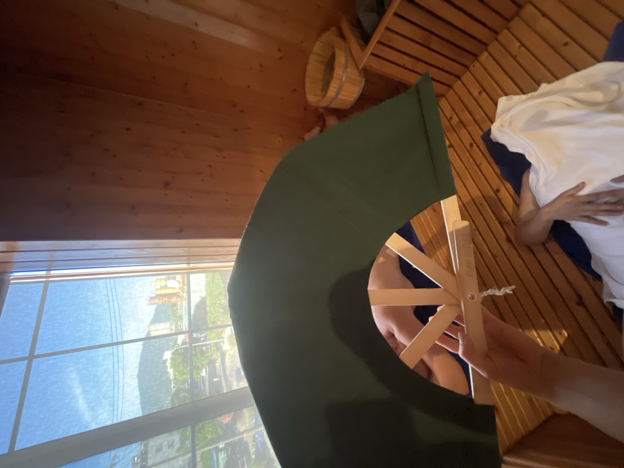 A first-person perspective shot within a wooden sauna. A dark green fan-like object held by a hand obstructs the view of a person lying down, who is covered with a white towel. The sauna features wooden benches and a wooden bucket in the corner. Through a large window, a view of Đà Nẵng, Vietnam, is visible, showing buildings, trees, and a mountain in the background. The sky is bright blue with some clouds.