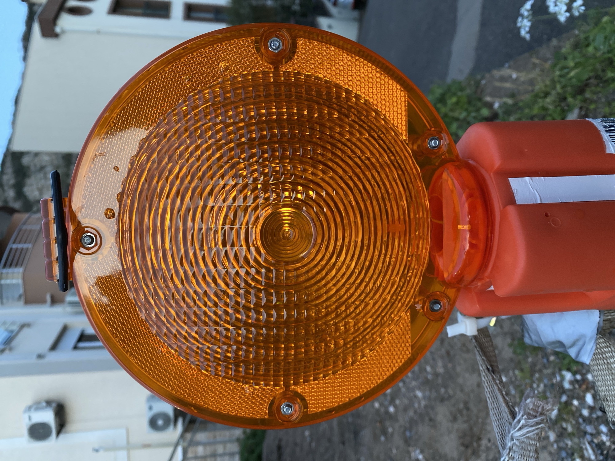 A close-up overhead shot depicts a round, orange construction warning light attached to a rectangular, orange plastic container. The light features concentric circles emanating from the center, with a crosshatch pattern around the outer edge. Silver screws are visible, securing the light's components. The container has white reflective tape adhered to it. The background shows a building facade with white walls and dark window frames, as well as some greenery and a glimpse of what appears to be a road or walkway. The location is possibly within an urban environment, and Budva, Montenegro, is named as the city, though the image itself does not provide explicit location cues.