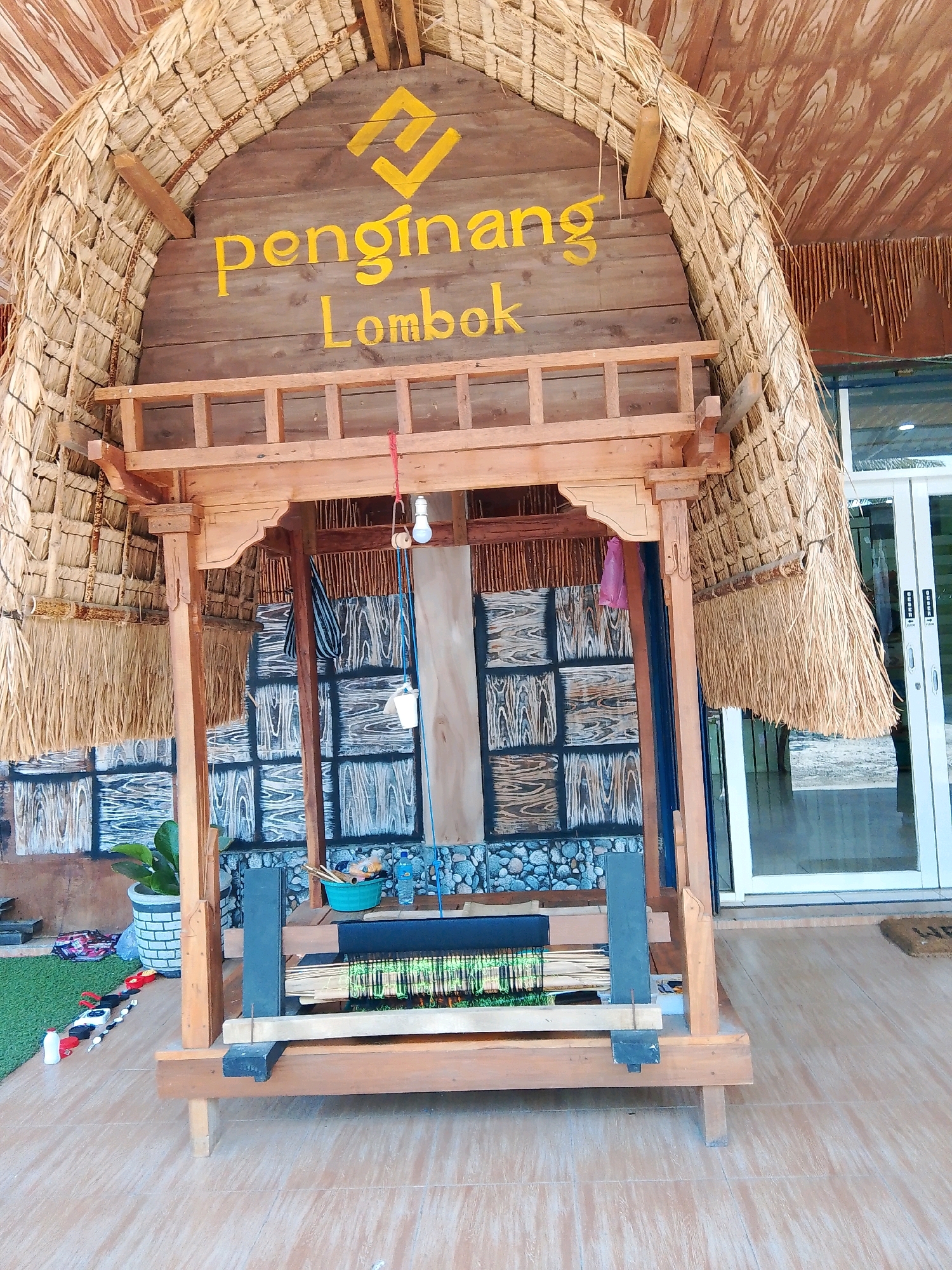 The image shows the entrance to a building with a thatched roof and wooden accents. A sign above the entrance reads "penginang Lombok" in yellow lettering, with a yellow symbol above it. Below the sign is a wooden structure with woven fabric or mats on display, and a weaving loom. To the left, there is a potted plant and some colorful items on a green mat. To the right, there are glass doors, suggesting the entrance leads into a shop or accommodation. The building appears to be in a tropical setting, possibly part of a resort or cultural center in Bunkate, Indonesia, given the mention of Lombok in the signage. There are no people visible in the image.
