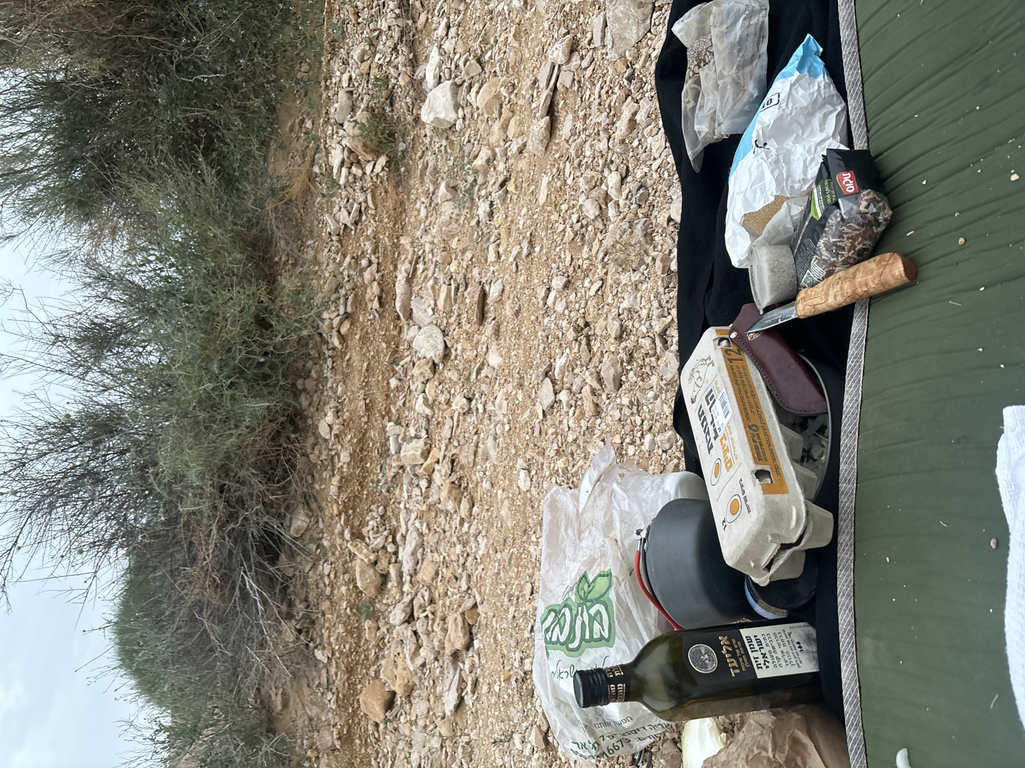 The image shows camping gear and food supplies arranged on a surface, seemingly outdoors. There is an olive oil bottle labeled "Eliad," a carton of eggs, and various packages of food. A knife is also present. The background suggests a rocky, desert-like environment, with sparse vegetation. Based on the text on the olive oil bottle "from the Golan Heights", the location is likely the Hevel Eilot Regional Council, Israel.