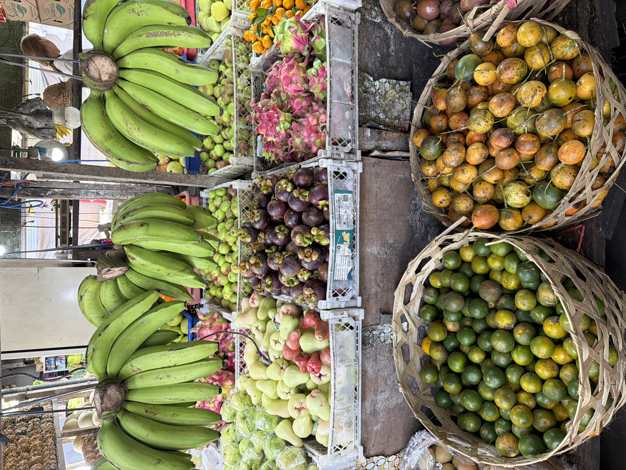 This is a photograph of a fruit stand in Ungasan, Indonesia. Bunches of green bananas hang from above. Below the bananas, fruits such as apples, dragon fruit, mangosteens, water apples, oranges, passion fruit, and limes are displayed in plastic crates and woven baskets. The fruits are stacked and arranged for sale, creating a colorful and abundant display.