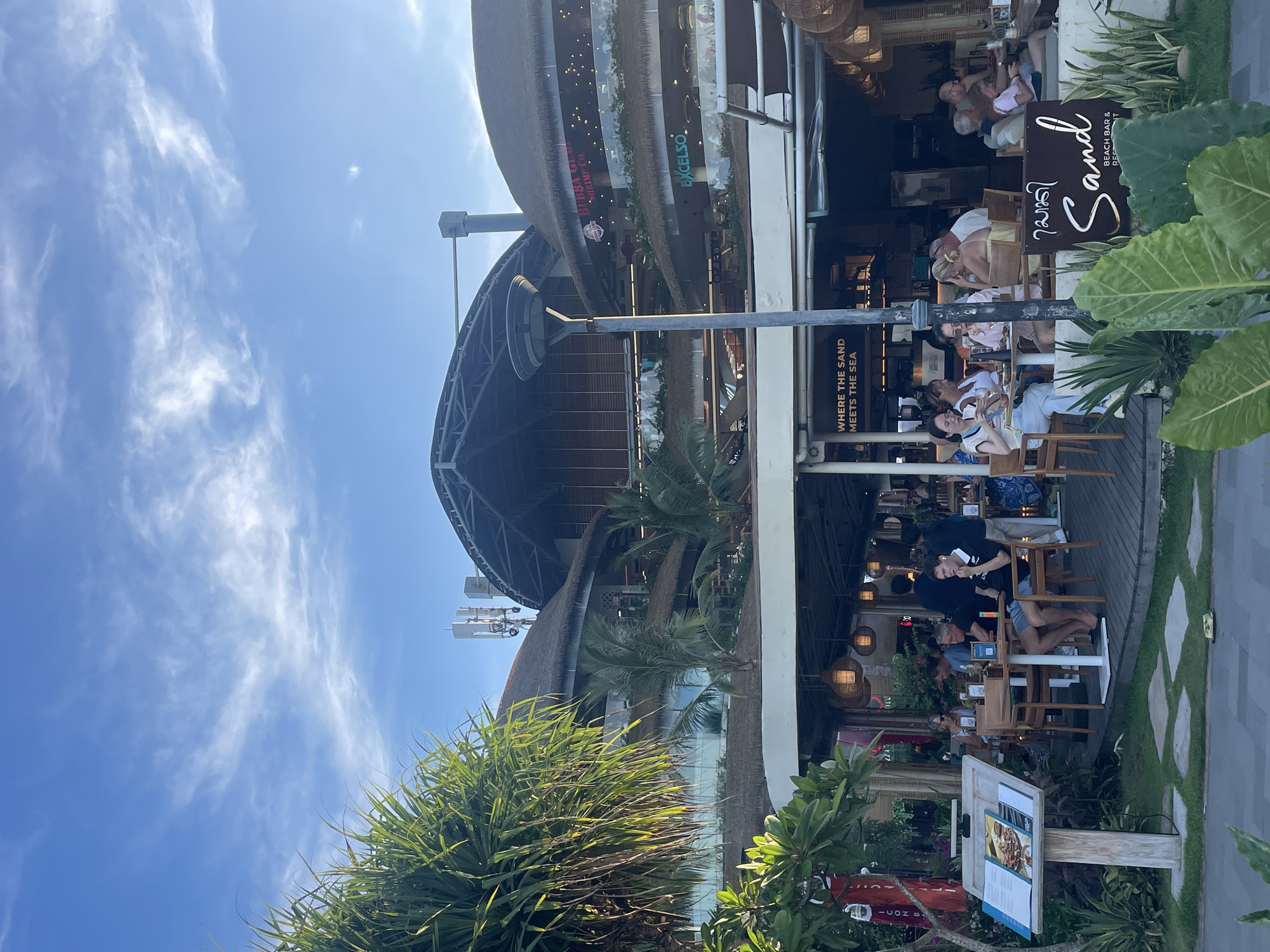 This image captures a vibrant, sunny daytime scene at a multi-level outdoor restaurant and bar complex in Denpasar, Indonesia. The setting features tropical architecture with distinctive thatched roofs and lush green foliage, including palm trees and other dense plants, framing the foreground and surrounding the establishment. The sky above is a clear, bright blue with scattered, wispy white clouds, indicating pleasant weather.
Numerous individuals are gathered at tables on various levels of the complex, suggesting a lively dining or social event. Patrons are seen casually dressed, engaged in conversation, eating, and drinking. The atmosphere appears relaxed and inviting.
Prominent signage identifies several businesses. A large wooden sign in the mid-ground clearly reads "Sand BEACH BAR & RESTAURANT," indicating the primary venue. Below this, a smaller sign declares "WHERE THE SAND MEETS THE SEA," reinforcing the beachside or coastal theme. On the upper levels of the building, other visible business names include "BUBBA GUMP SHRIMP CO" and "EXCELSO," suggesting a variety of culinary options within the complex. A menu stand is also visible in the lower foreground.
The scene conveys a sense of leisure and enjoyment in a resort-like environment. The bright natural light, the tropical greenery, and the architectural style all contribute to the impression of a popular vacation or leisure spot.