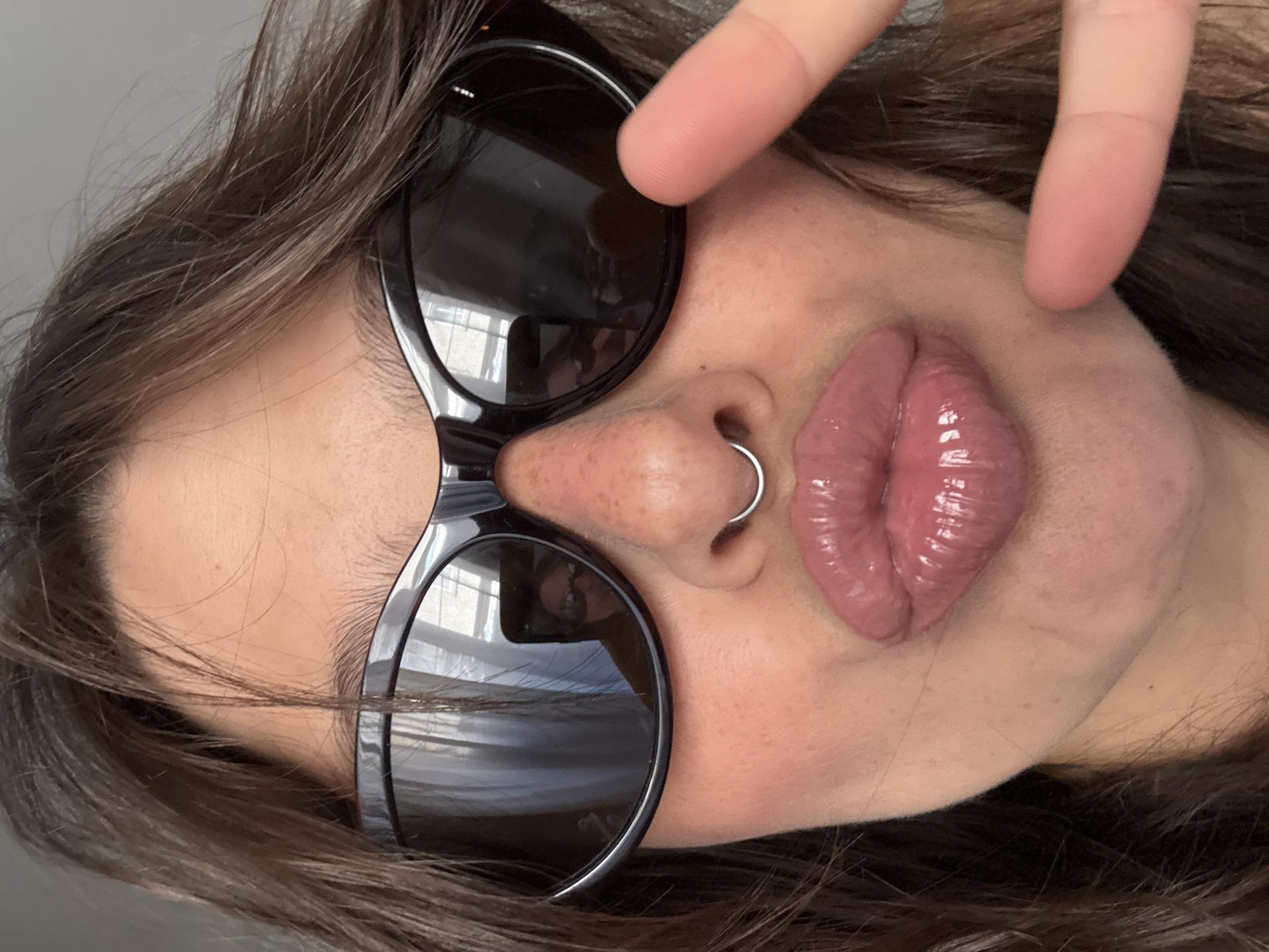 A close-up, tilted portrait shows a woman with brown hair wearing sunglasses and a nose ring. She is holding a finger near her temple. Reflections in the sunglasses indicate an interior space with light coming through a window. There are no clear indicators in the image to determine a specific location in Tel Aviv, Israel.
