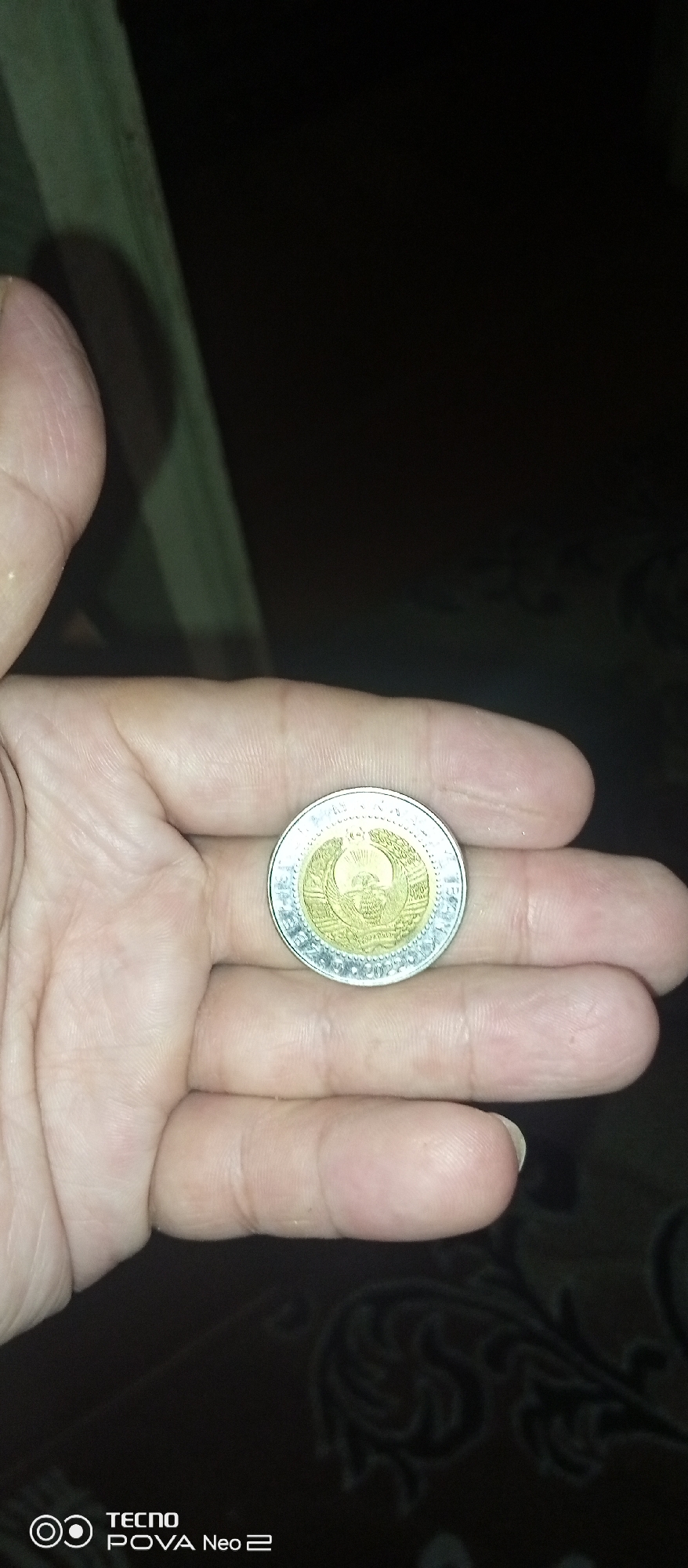 The image shows a close-up of a hand holding a coin. The coin has a central golden emblem surrounded by a silver rim. The emblem appears to be a crest with a sunburst and a bird. Text is visible around the rim, but it is not clearly legible. The hand is light-skinned and appears to be resting on a dark, patterned surface. The background is dark and out of focus, with a green object partially visible on the left side. The scene is indoors, and the lighting suggests it is nighttime or a dimly lit environment. The resolution of the image is moderate, with some graininess visible. There is no indication of the city of Tahmach, Uzbekistan.