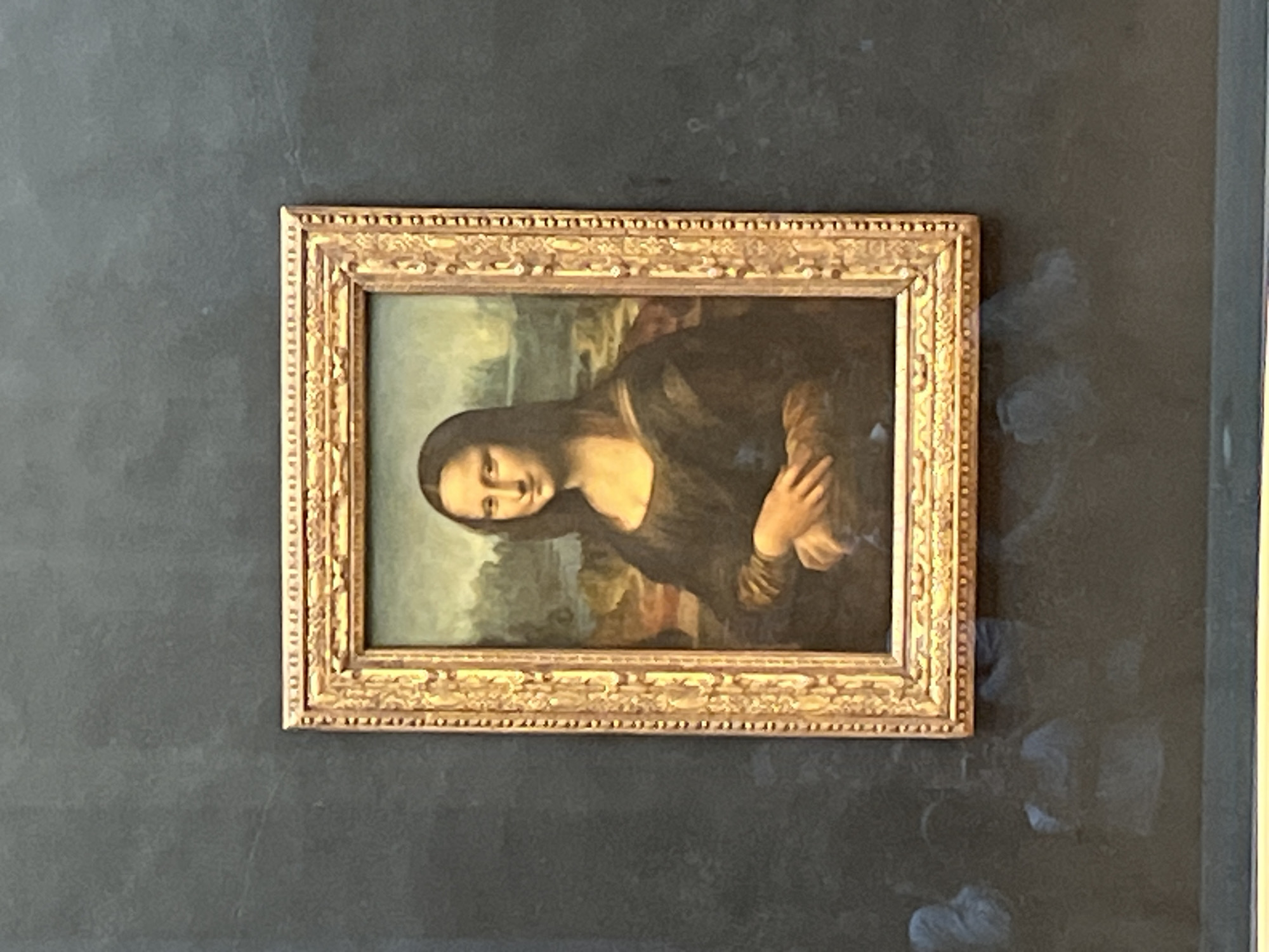 The image shows the Mona Lisa painting, encased in a gilded frame, hanging on a wall that appears dark grey or black. The painting is displayed behind protective glass, which reflects the glare of the room. Given the context, this image was likely taken in the Louvre Museum in Paris, France, where the Mona Lisa is permanently exhibited.