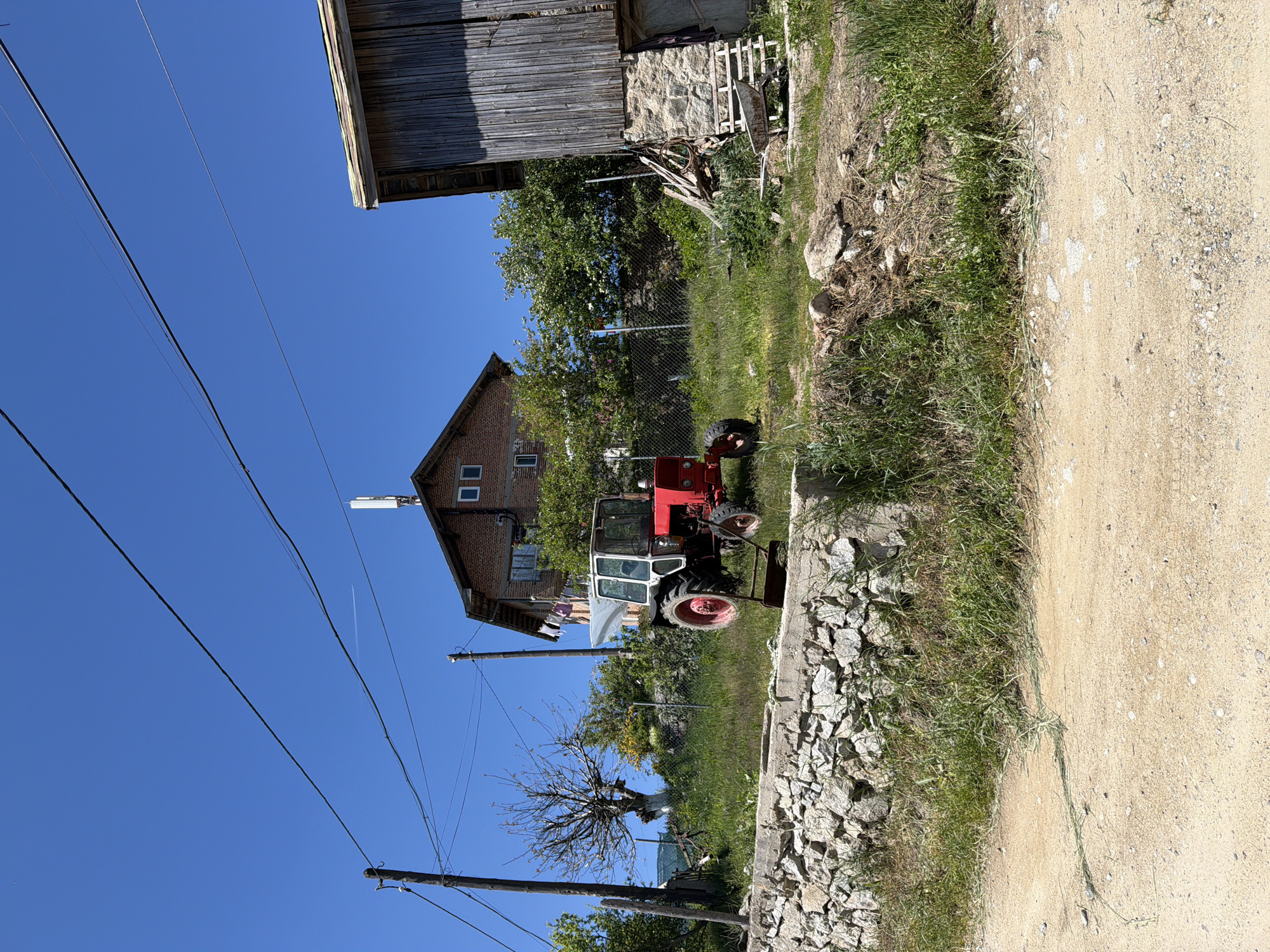 The image depicts a rural scene, possibly in Avramovo, Bulgaria. A red and white tractor is parked on a grassy area next to a stone wall and a dirt road. A brick building with windows and what appears to be a satellite dish on the roof is partially visible. A wooden structure, possibly a barn, is at the top of the image. Power lines run across the blue sky. A bare tree and other vegetation are scattered throughout the scene.