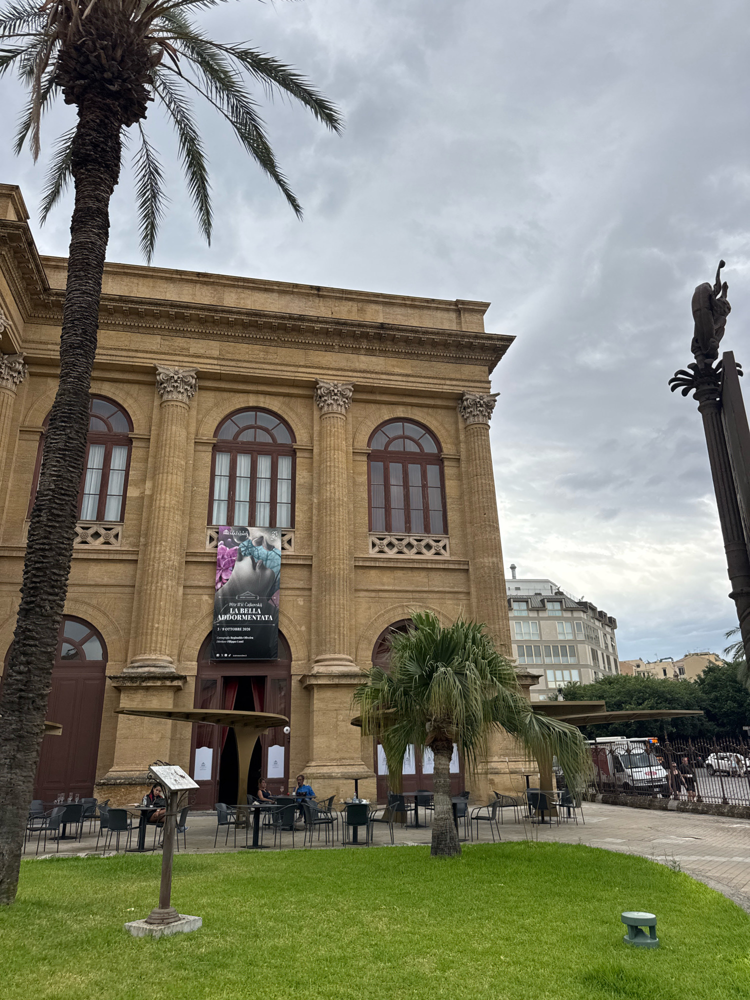 This image depicts the Teatro Massimo in Palermo, Italy, on an overcast day. A large palm tree is positioned to the left of the frame, with its fronds extending upwards. The theater itself is a grand, tan-colored building with classical architectural elements, including columns and arched windows. A banner hangs in the center of the facade, advertising an event titled "LA BELLA ADDORMENTATA" with a date of "3/OTTOBRE 2020". In front of the theater, an outdoor seating area with tables and chairs is set up, some with umbrellas providing shade. Several people are seated at the tables, engaged in conversation or appearing to dine. A grassy lawn with a low-lying green light fixture is in the foreground. To the right of the theater, a tall monument with sculpted figures is visible, along with a street with cars and pedestrians beyond a metal fence. Modern buildings and trees are visible in the background, suggesting an urban setting.