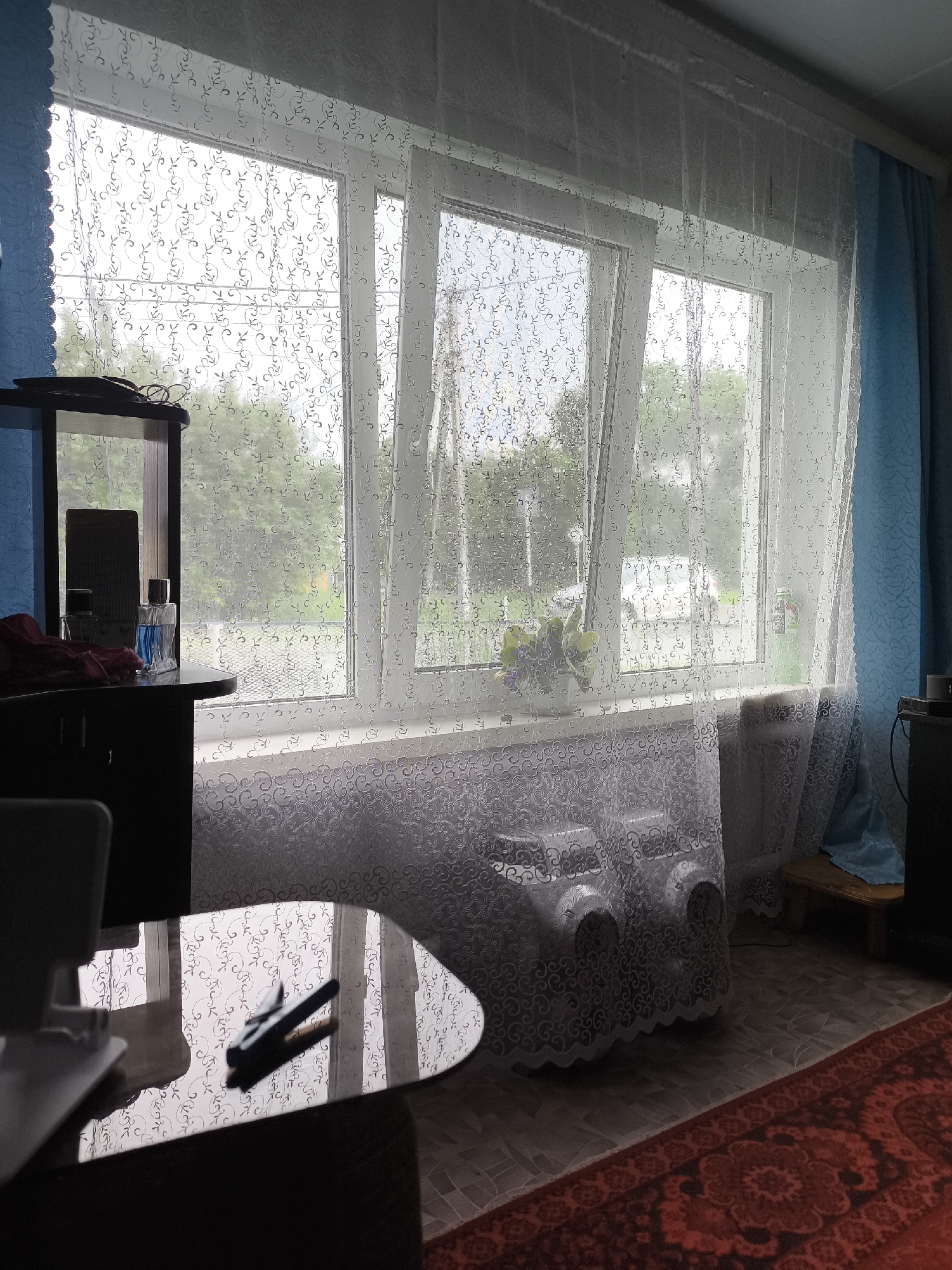 The image depicts an interior view of a room in Saraktash, Russia, likely a living space or bedroom, looking out through a window. The window is covered with a sheer white curtain featuring a floral pattern, and behind it, a blue curtain is visible on the right. Outside the window, trees and a cloudy sky are discernible.
In the foreground, to the left, a dark, possibly wooden, cabinet or dresser is partially visible, with a mirror reflecting the window. On top of the cabinet, there are two glass bottles, one appearing to contain a blue liquid, and a red piece of fabric. A dark, reflective table with a curved edge occupies the lower portion of the image, with a stapler resting on its surface. The reflection on the table shows the patterned curtain and the window.
The floor is covered with a patterned carpet in shades of red and dark brown, with linoleum visible in the area beneath the window. The windowsill is covered by a patterned fabric, and two white objects, possibly shoe covers or decorative items, are placed on it. A radiator is visible beneath the windowsill, also covered by the patterned fabric.
The lighting suggests an overcast day. No people are present in the image. The environment appears to be a residential setting in a Russian city.
