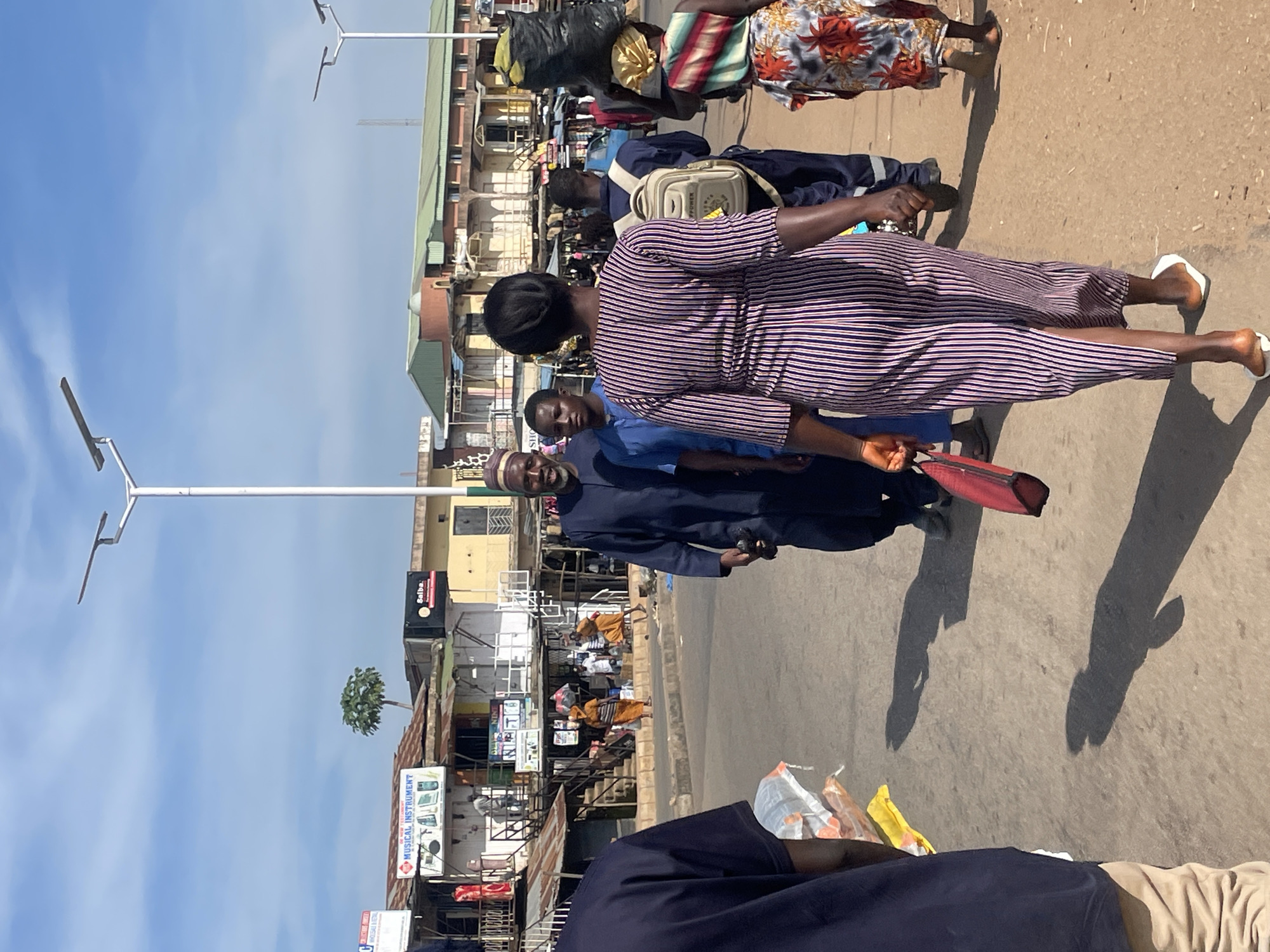 In Jos, Nigeria, a busy street scene unfolds under a bright, partly cloudy sky. The image captures a group of people walking along a paved road, with buildings and market stalls lining the background. The foreground is dominated by the backs of several individuals. A woman in a purple and white striped dress with a high slit walks away from the camera, carrying a red bag in her left hand and wearing white sandals. To her left, a man in a dark blue shirt and khaki pants is partially visible. Behind her, a person in a blue uniform with a light-colored backpack walks, and further back, a woman with a headwrap carries a large black bag of goods on her head.
The middle ground shows several men walking towards the camera. One man, wearing a dark blue robe and a patterned cap, smiles at the camera and holds a black object in his right hand. Beside him, a young man in a light blue outfit also walks. In the background, market stalls with various goods are set up. One stall displays signs for "MUSICAL INSTRUMENT" and another for "Salba." Other structures include buildings with green roofs and colorful facades. Streetlights are visible in the distance, suggesting this is a commercial area. The ground is dusty asphalt, and shadows are cast by the sun, indicating it is daytime.