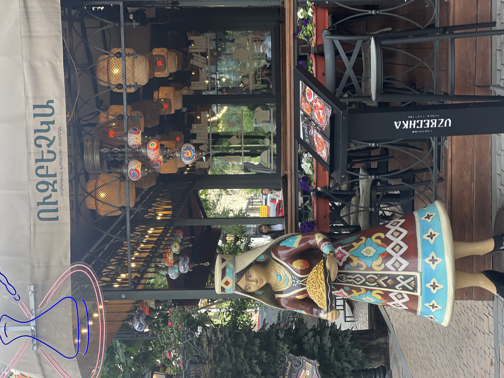 A street view in Yerevan, Armenia, shows a restaurant facade. The restaurant, "Ubeechka," features an awning in Armenian text, decorative lanterns, and a sign with menu items. A colorful statue of a woman in traditional clothing holding a dish stands at the entrance. Outdoor seating is visible. The scene includes greenery and pedestrians in the background.