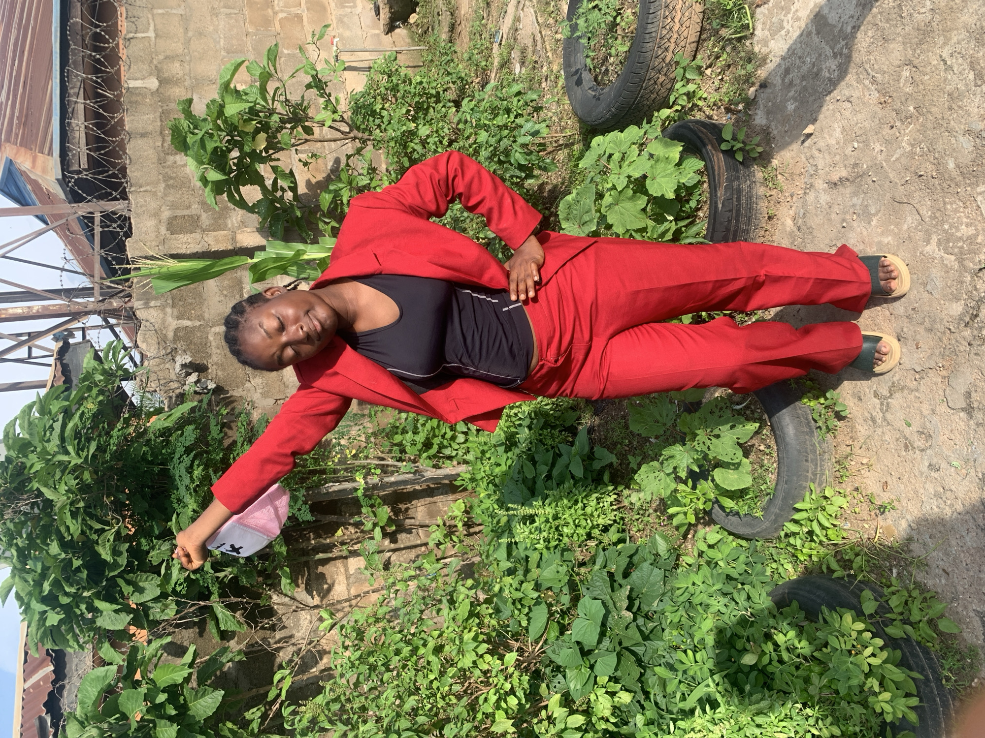 A young Black woman stands outdoors in Sabon Tasha, Nigeria, dressed in a red suit and black top. She holds a pink and white cap in her left hand, which is raised above her head. Her right hand rests on her hip, and she gazes downwards with her eyes closed. The background consists of a brick wall, lush green vegetation, and discarded tires used as planters. The overall scene suggests a casual outdoor setting, possibly a yard or garden area within the city.