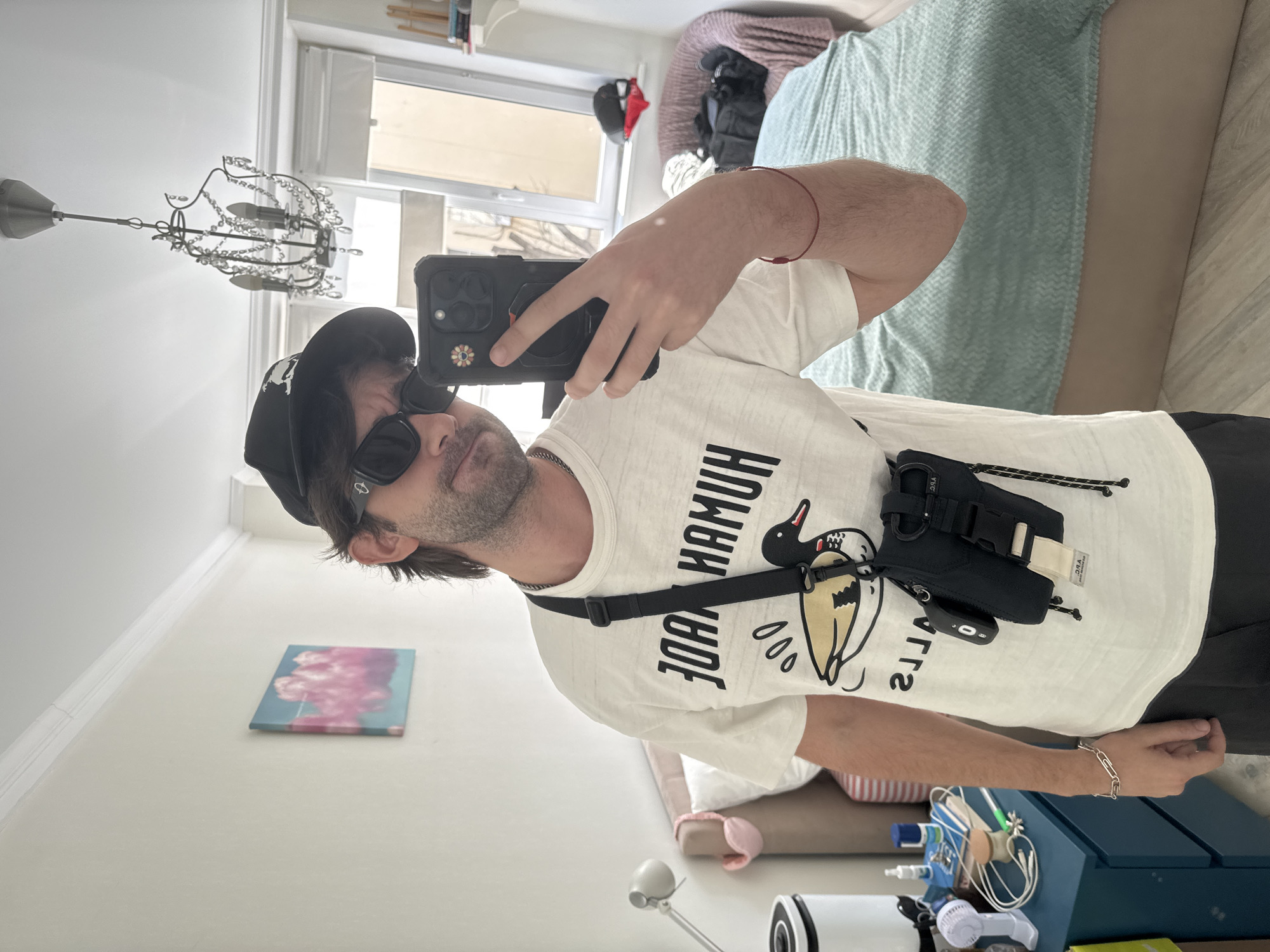 A man is taking a selfie in a bedroom. He is wearing a white t-shirt with a duck graphic and text that reads "JOAN HAMUN", a black baseball cap, and sunglasses. He has a black crossbody bag with multiple compartments hanging from his neck. In his right hand, he is holding a smartphone. On his left wrist, he wears a silver chain bracelet, and on his right wrist, a red string bracelet. The background shows a bed with a mint green blanket, a bedside table with a lamp and personal items, a window, and a chandelier hanging from the ceiling. A small painting of pink clouds is visible on the wall. The room appears to be a personal living space. The overall lighting suggests it is daytime.