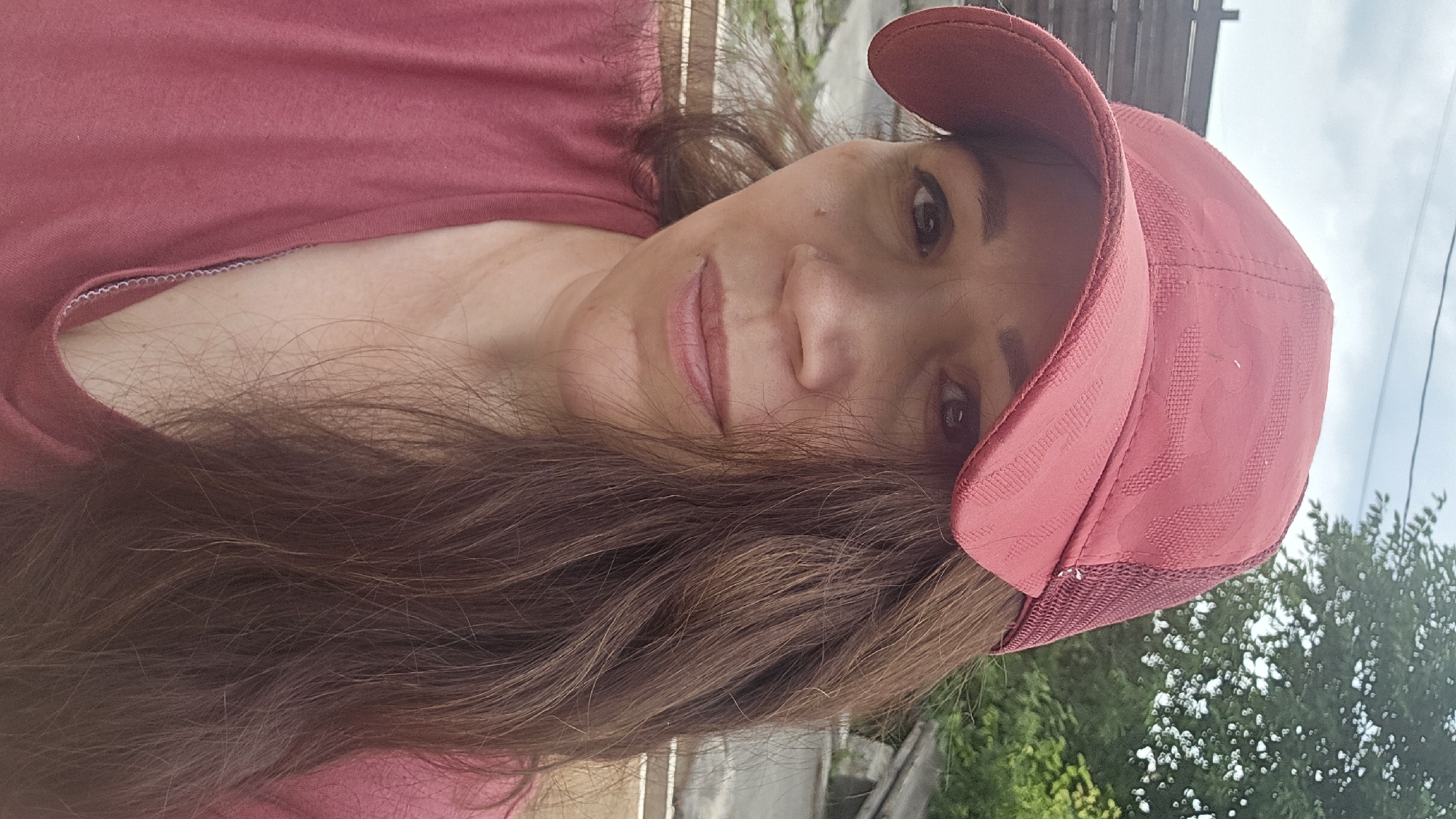 A medium-shot, outdoor, close-up selfie captures a woman wearing a red baseball cap and a pinkish-red t-shirt. She has long, brown hair that partially obscures her face. Her eyes are brown, and her lips are slightly parted. The background suggests an outdoor setting, possibly a yard or street, with a blurred out wooden fence, some greenery, and a cloudy sky. The overall lighting is natural, indicating it's daytime. The image is taken from a slightly low angle.
