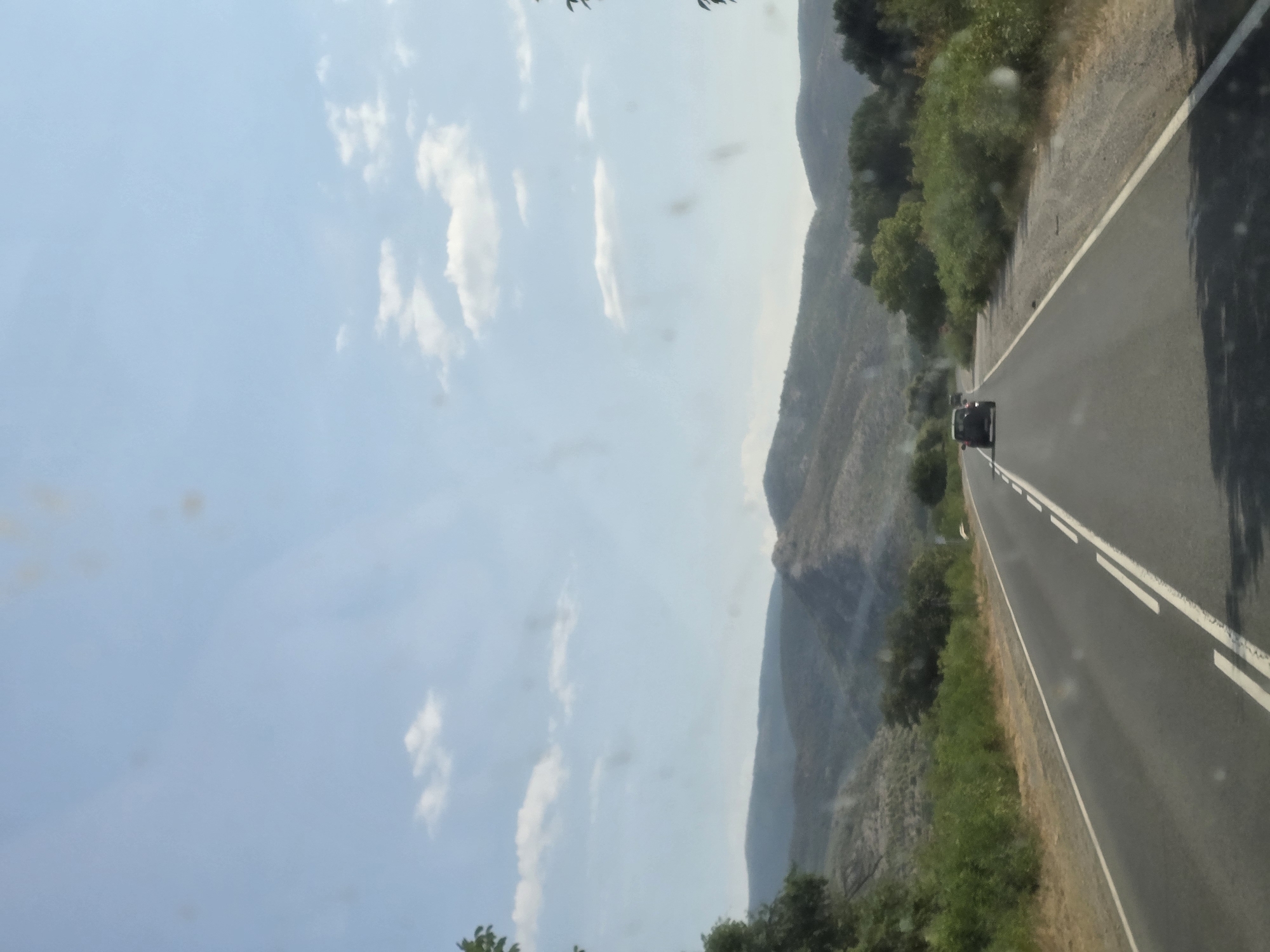 The image depicts a road traveling through a hilly, sparsely vegetated landscape under a clear blue sky with scattered white clouds. The road is paved and has white lane markings. In the distance, there are two vehicles, one in front of the other, traveling in the same direction. The foreground shows the road surface and the edge of the road lined with dry grass and bushes. The background consists of rolling hills covered in scrub and distant mountains. The scene appears to be captured from inside a moving vehicle. The image is from Binkos, Bulgaria.