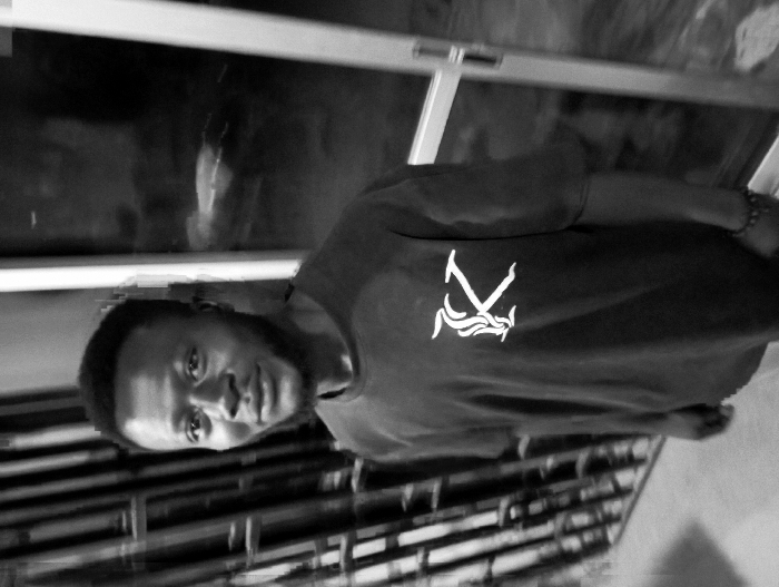 The black and white image displays a man posing for a photograph in an indoor setting. The image is oriented vertically, with the man's head positioned towards the left side of the frame. If rotated 90 degrees counter-clockwise to a standard upright orientation, the man would be standing facing slightly to the viewer's left.
The subject is an adult male with short, dark hair and a neatly trimmed beard or stubble. He is looking directly at the camera with a neutral to slightly engaged expression. He is wearing a dark, short-sleeved t-shirt. On the front of his t-shirt, a distinctive white logo is visible. The logo appears to be a stylized capital letter "K", where the diagonal and vertical strokes on the right side of the 'K' are replaced or embellished with multiple horizontal, wavy, flame-like, or fluid lines.
The background is somewhat blurred but reveals an indoor environment. Behind the man's upper body, there appears to be a window or a glass partition with reflections, and possibly a glimpse of an outdoor or dimly lit area beyond. Behind his lower body and to his right (when viewed upright), there are horizontal elements that resemble blinds, slats, or a railing, suggesting part of a room's interior design. The lighting is moderate, creating some shadows and highlights that emphasize the man's features and the texture of his shirt. The overall mood of the image is casual and direct.