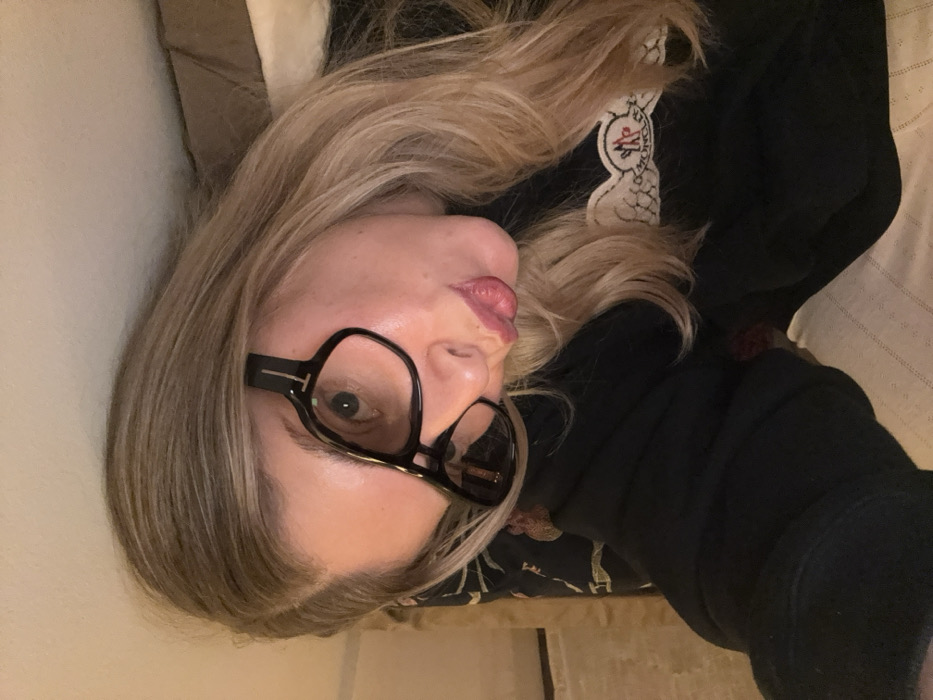 A woman with long blonde hair and glasses poses for a selfie. She is wearing a black hoodie with a white logo on the collar. She is lying on her side, with her head tilted towards the camera. The background is a mix of neutral tones, suggesting a bed or couch setting. She has light skin and a slight pout.