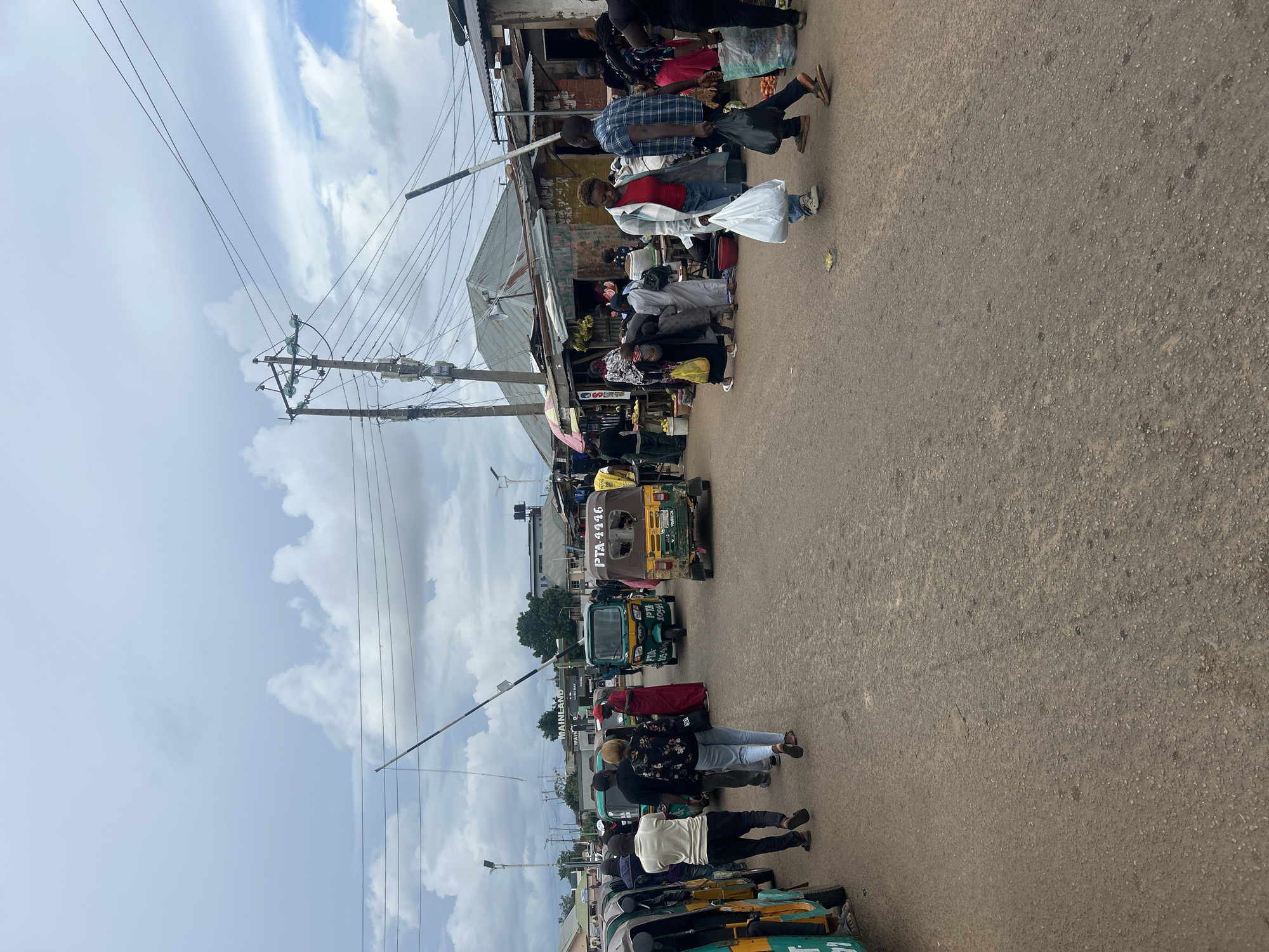 The image depicts a busy street scene in the city of Jos, Nigeria, likely near a market area. The foreground is dominated by a dusty, paved road with several auto-rickshaws, also known as "keke naphap," parked or moving. People are walking on the road and sidewalks, some carrying shopping bags.
In the background, stalls and small shops line the street, constructed from corrugated metal and wood. There are signs visible, including one that reads "MAINLAND." Overhead, power lines and poles are prominent against a cloudy sky. The overall impression is one of a vibrant, bustling urban environment, typical of many Nigerian cities.
The scene includes various individuals engaged in daily activities. Some are interacting with vendors, while others are simply moving through the area. The clothing of the people suggests a warm climate. The presence of the auto-rickshaws indicates a common mode of transportation in the region. The architecture of the buildings, with their makeshift nature, is characteristic of informal markets and commercial areas in Nigeria. The ground is rough and uneven, with visible debris.