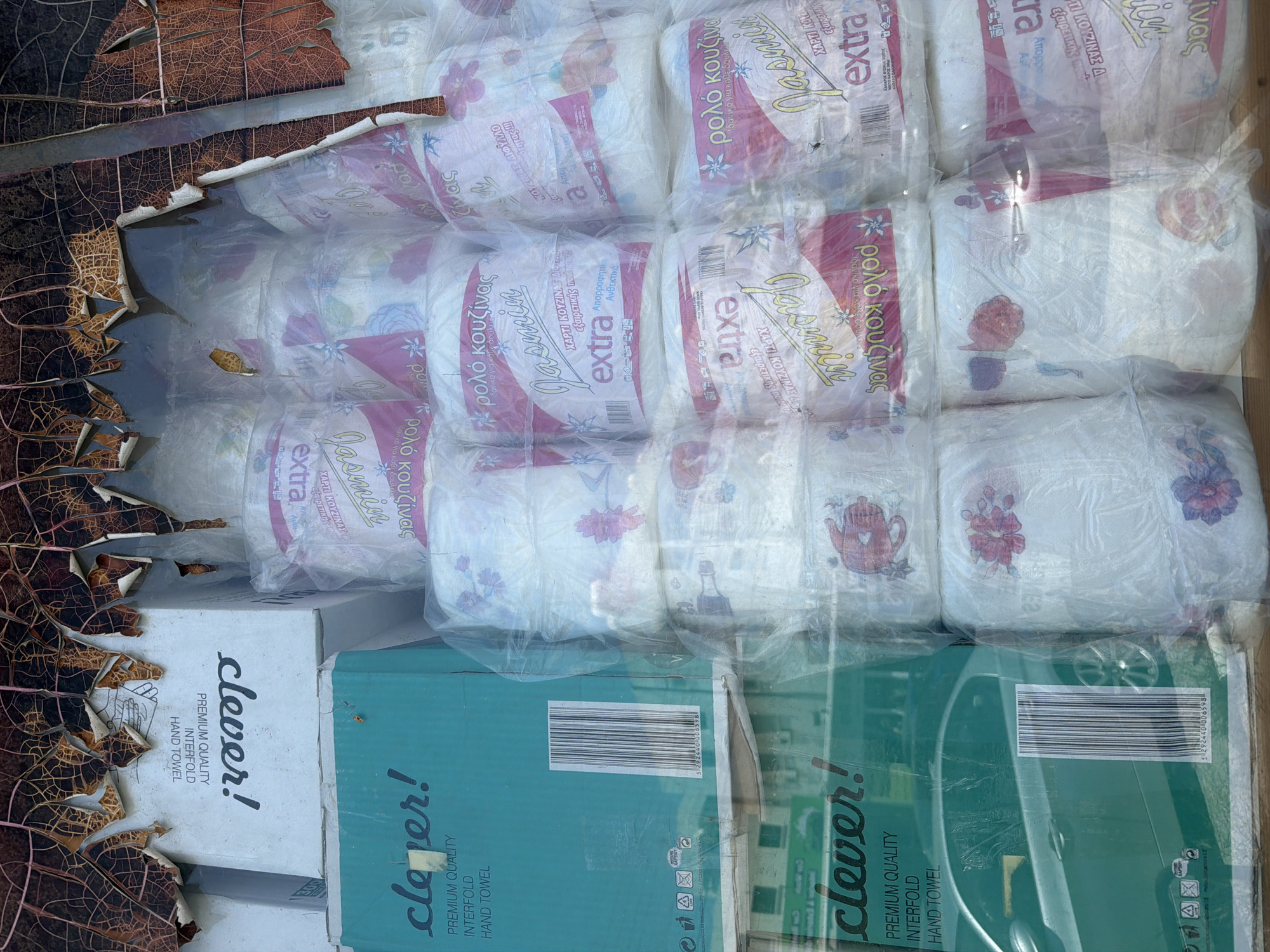 The image shows a close-up view of stacked toilet paper rolls and boxes of hand towels outside a shop in Larnaca, Cyprus. The products are packaged in plastic and cardboard, with branding that includes "clever!" and "Jasmine extra." Some of the packaging is in Greek. The scene is partially obscured by torn, brown, leaf-like material, suggesting it might be an outdoor display with some weathering. The background appears to be the facade of a building with reflections of a car on the "clever!" hand towel box. The overall impression is of merchandise displayed for sale in a somewhat disorganized or weathered outdoor setting.