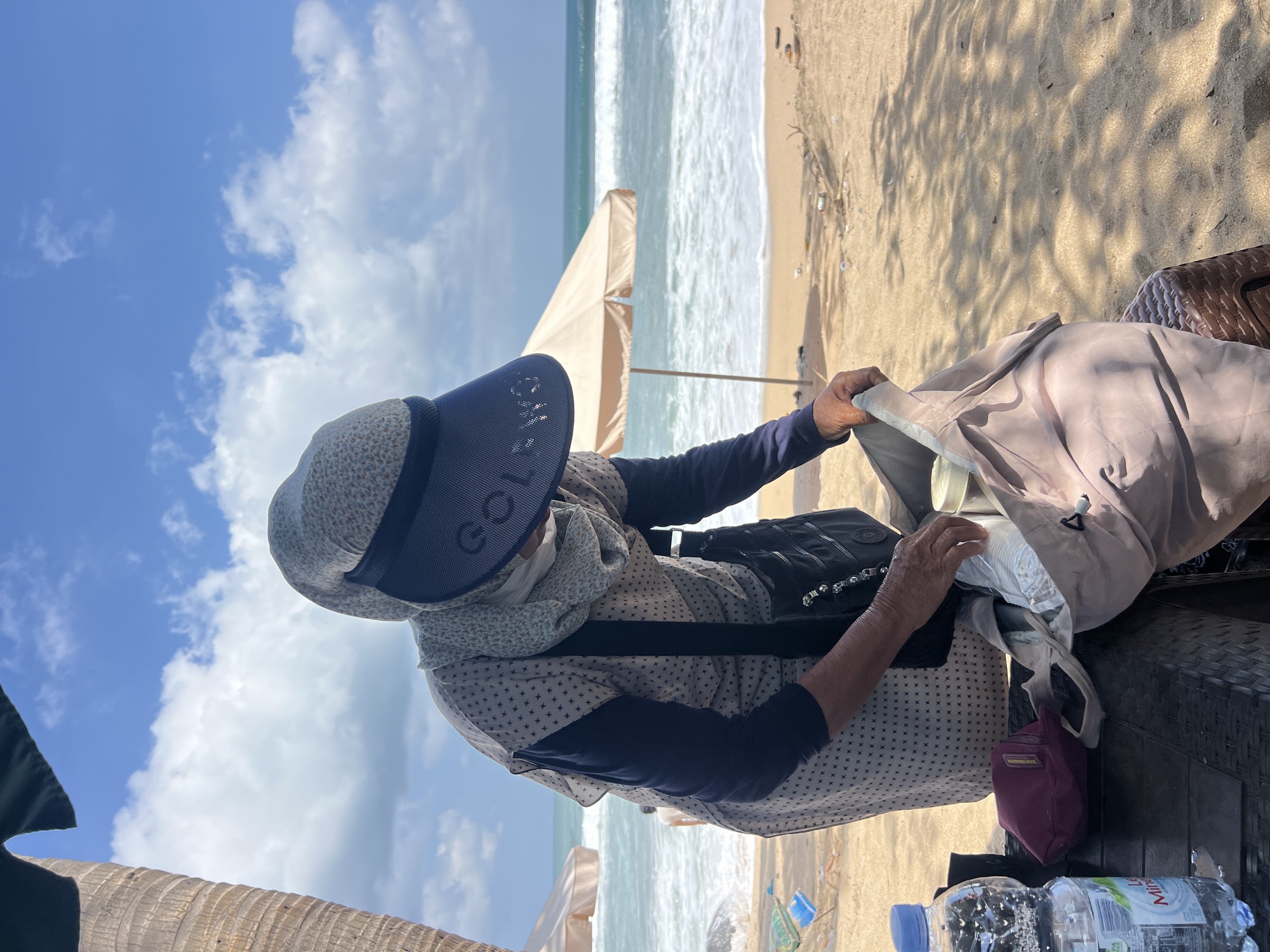 A person wearing a hijab, a patterned face mask, a light-colored patterned shirt with dark sleeves, and a dark visor with "GOLFEWO" printed on it is standing on a beach in Kuta, Indonesia. The person is reaching into a light-brown fabric bag, which is open and contains some items. To the left of the bag, there is a maroon pouch and a clear plastic bottle of water on a dark, woven surface. The background shows a sandy beach with scattered litter, two beige beach umbrellas, and the ocean with waves under a blue sky with white clouds. There is a palm tree trunk on the far left of the image.