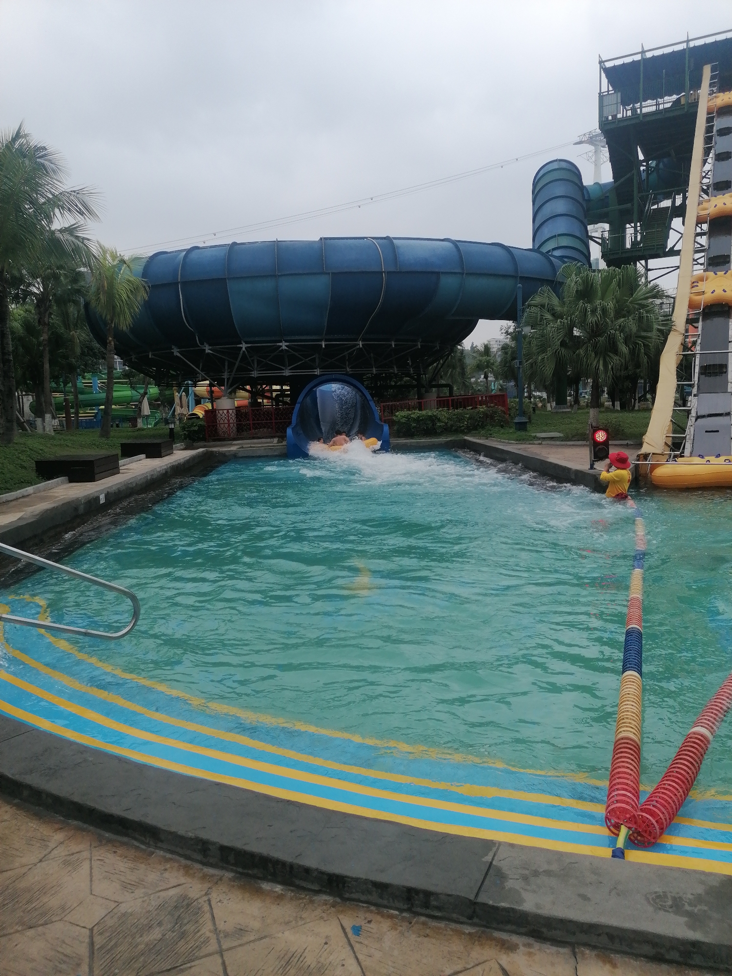 The image depicts a water park scene in Bai Chay Ward, Vietnam. A large blue waterslide structure dominates the background, emptying into a pool. People are visible emerging from the slide. A lifeguard in red attire is positioned near the edge of the pool next to a line of buoys. The pool has blue and yellow striped markings along the edge. Palm trees and other water park structures are visible in the background. The sky is overcast.