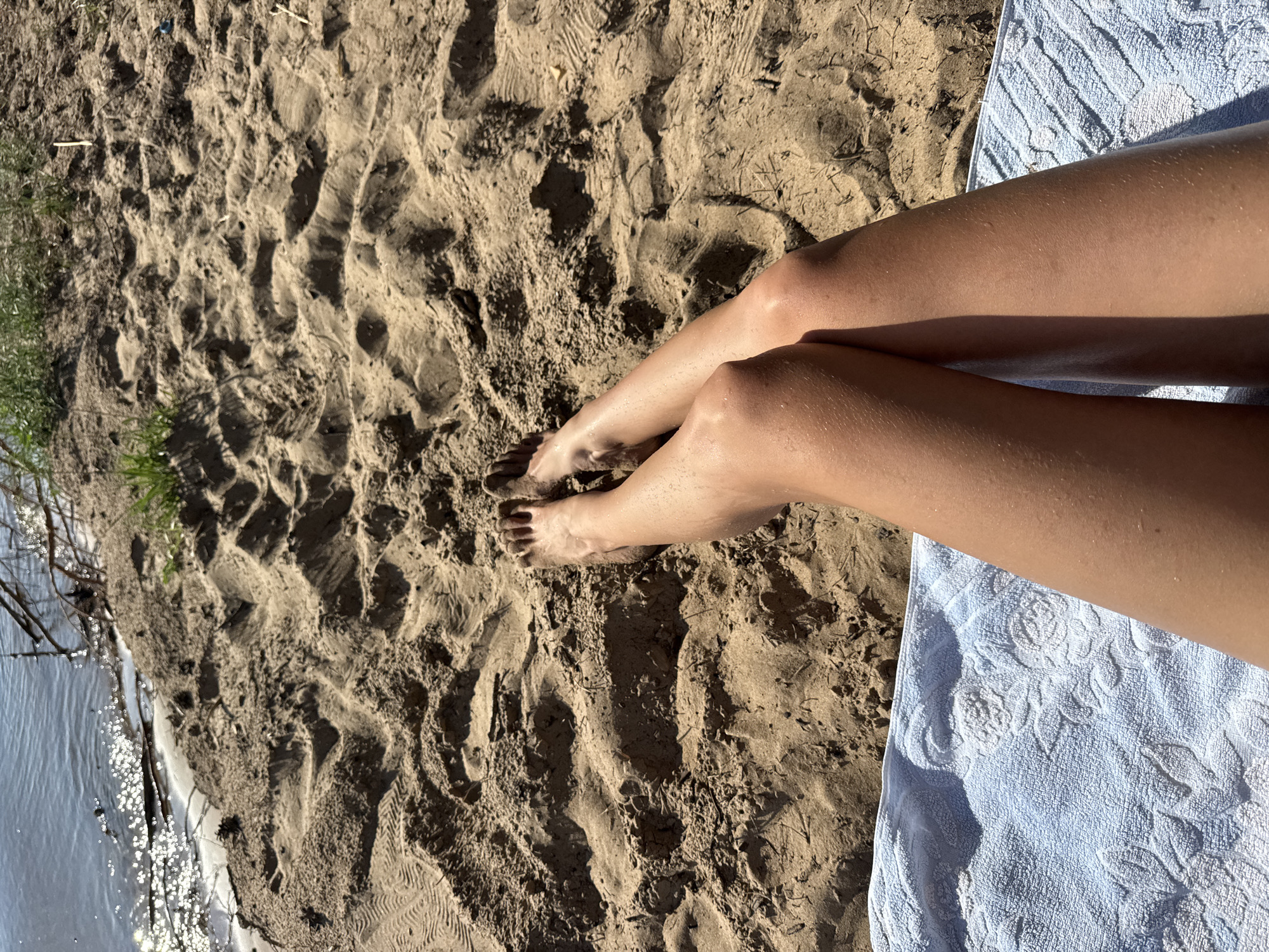 The image shows a person's legs lying on a sandy beach next to a body of water. The sand is light brown and textured with footprints and small debris. The water is calm and reflects sunlight. A light blue towel with a floral pattern is partially visible on the right side of the frame, underneath the person's legs. The lighting suggests it is daytime, likely sunny, given the shadows cast by the footprints and the sparkle on the water. The scene evokes a sense of relaxation and outdoor leisure, possibly a summer day at the beach. There is no visible text in the image.