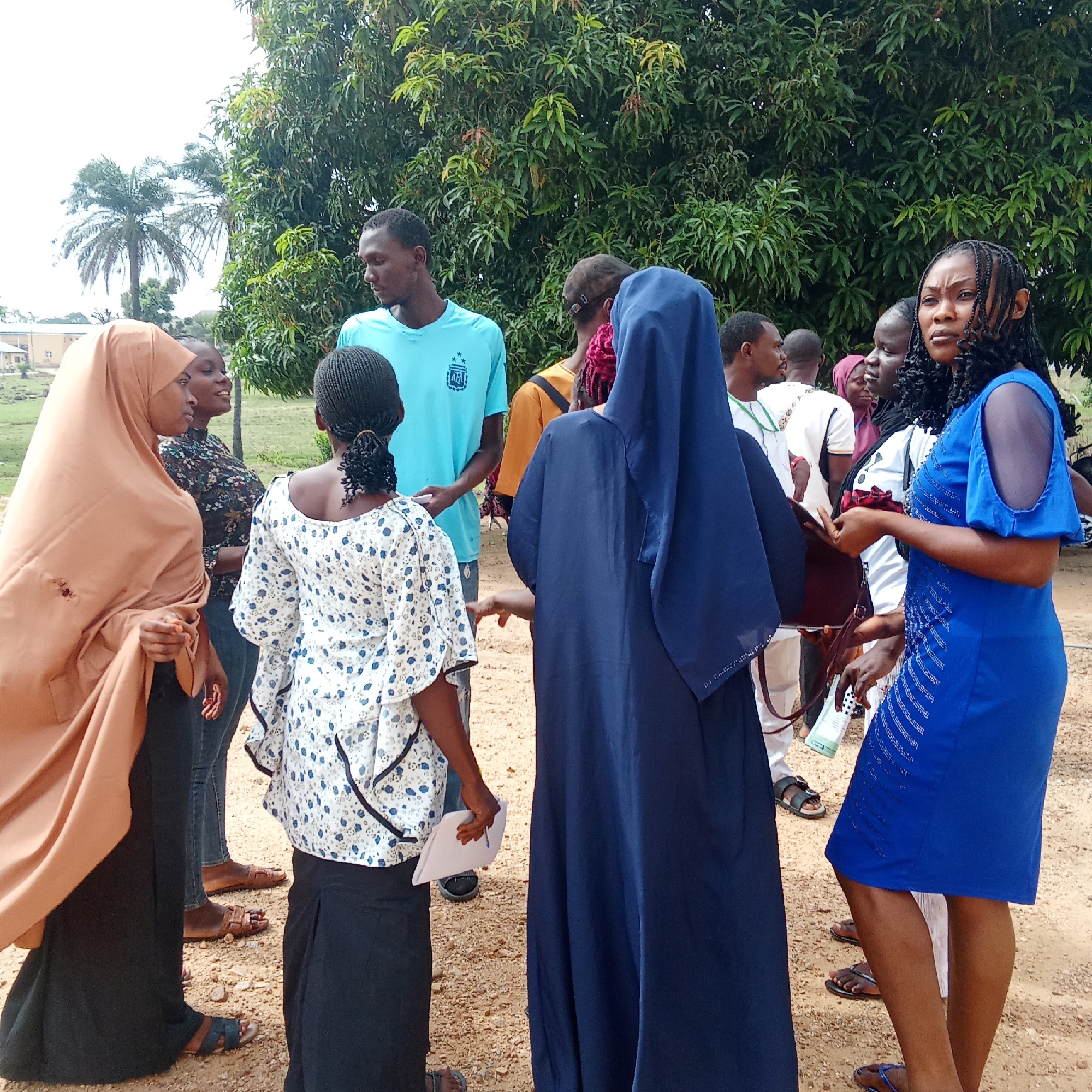 A group of people stand in a circle on a dirt ground in Akwanga, Nigeria. There are ten individuals visible, a mix of men and women. The women are wearing dresses and hijabs or modest religious attire. One woman in a bright blue dress looks to her right, holding a small bag in her hand. A woman in a tan hijab looks at another woman in a floral blouse and black skirt. Another woman is wearing a dark blue nun's habit. A man in a blue t-shirt stands in the background, while other men are seen behind the women in various light-colored shirts. Lush green trees dominate the background, with a palm tree and building visible in the distance to the left.