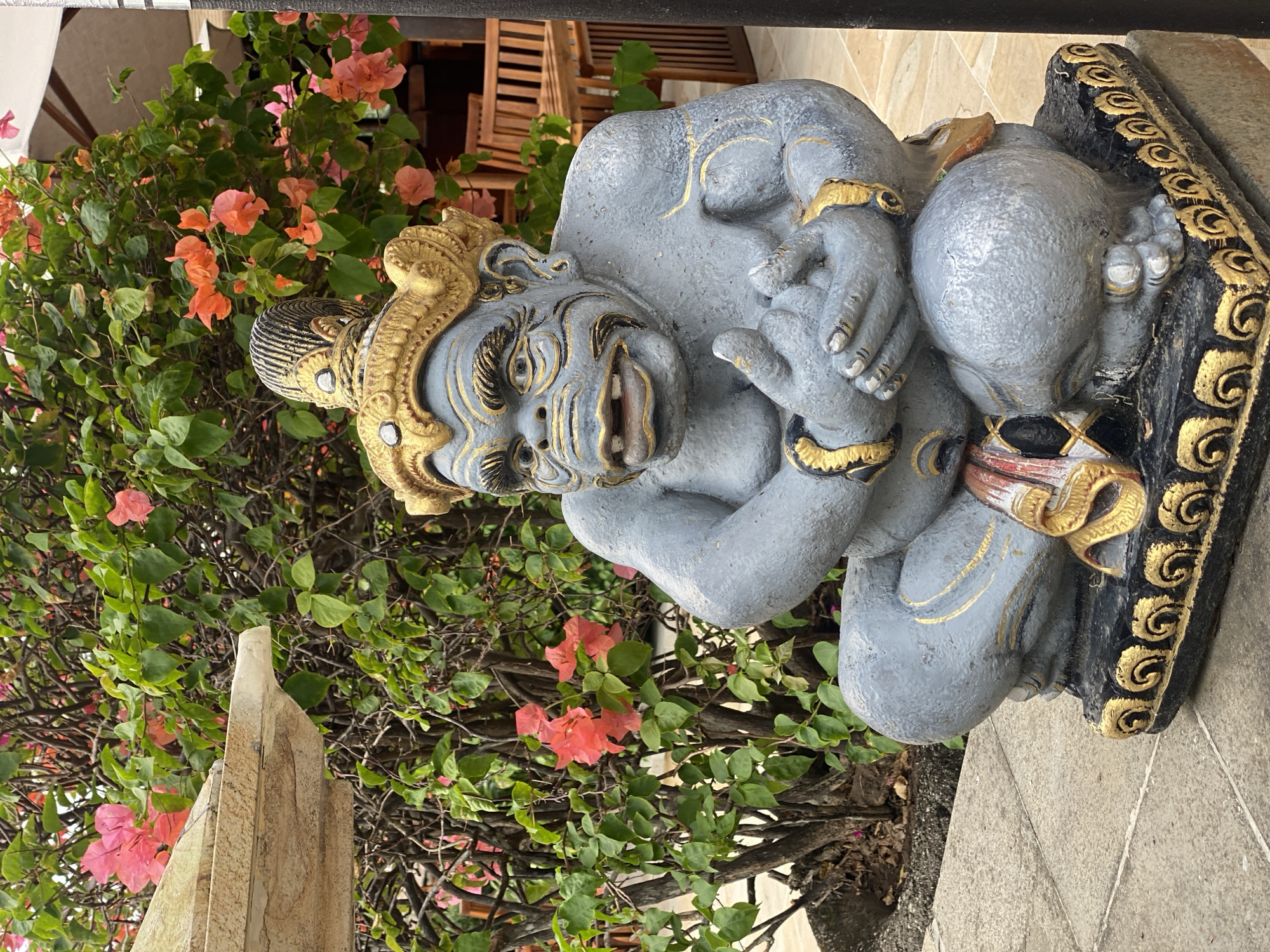 A gray stone statue of a mythical creature with a gold crown and gold accents sits in front of a lush green bush with pink and orange flowers. The creature is in a seated position, with its hands clasped together. The statue is placed on a black and gold decorative base, which rests on a paved walkway. In the background, wooden chairs and tables are visible, along with a white umbrella, suggesting an outdoor dining area. The overall scene evokes a tropical ambiance, possibly at a resort or restaurant in Kuta Selatan, Indonesia.