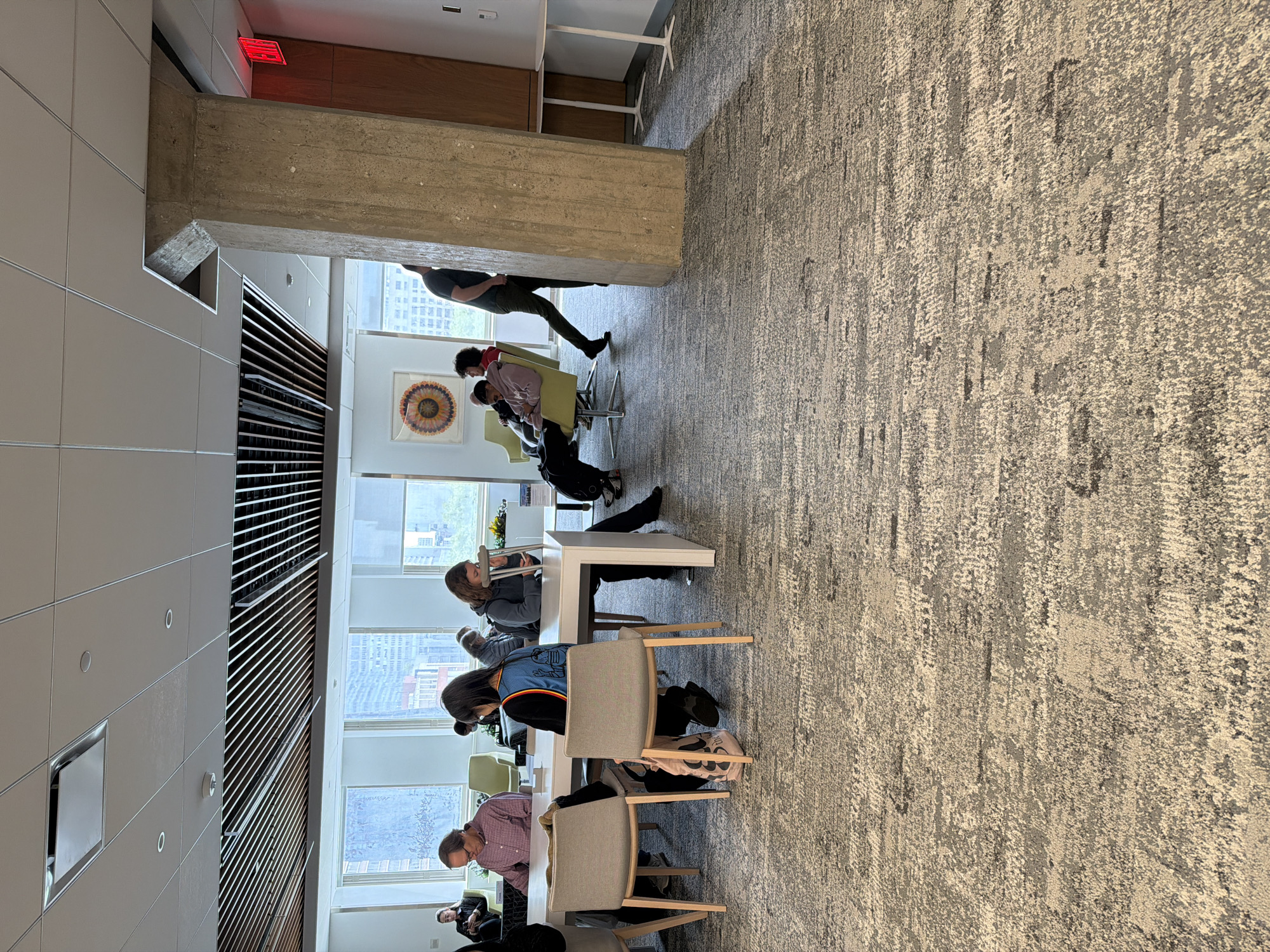 The image shows an indoor scene in what appears to be a modern office space. Several people are seated at a long white table, engaging in work or study. The room is well-lit with natural light coming through large windows, which offer a view of buildings, suggesting an urban location. One man is standing walking past the others near a concrete support beam. The presence of skyscrapers in the background suggests the location is likely in New York City, USA. There are several chairs, a decorative piece on the wall, and a textured grey and beige carpet. A grid of dark blinds or ventilation slats extends across the ceiling.