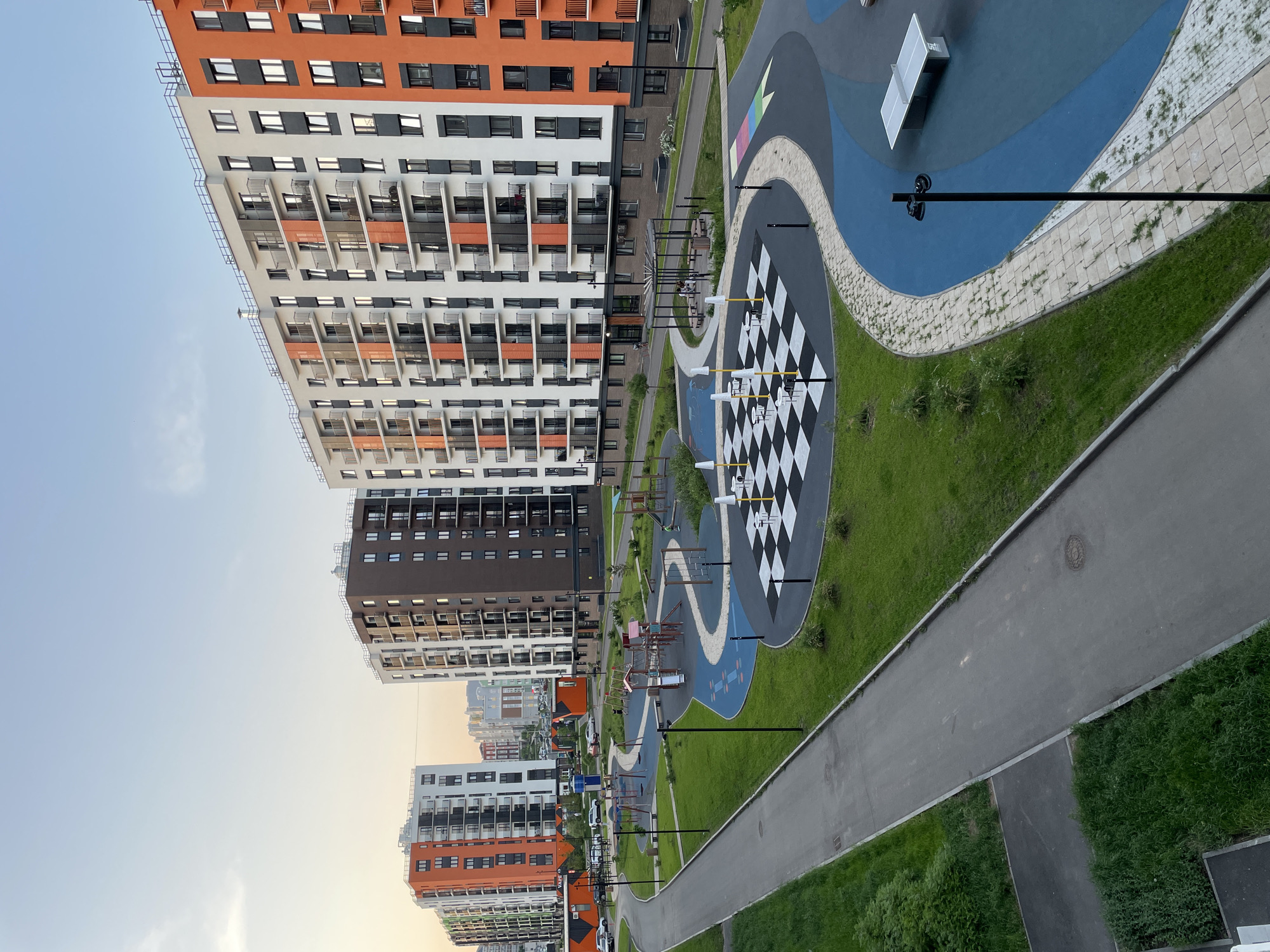 The image shows a playground area in Pervomaysky, Russia, nestled between tall apartment buildings. The playground features a large, painted chess board with oversized chess pieces, a play area with colorful surfaces, and a walking path. The buildings are modern with alternating orange, white, and brown facades. Several light poles stand on the playground.