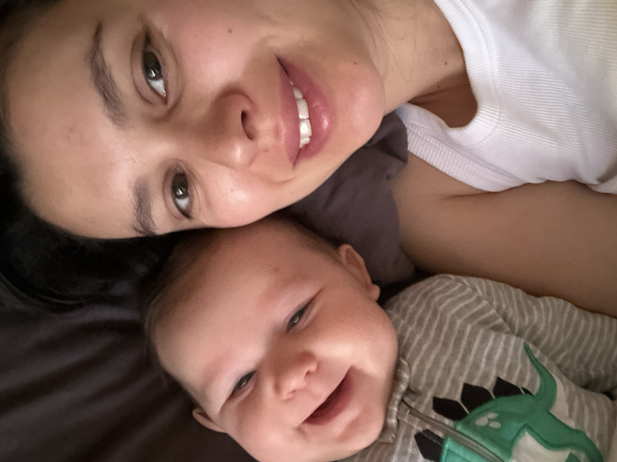 A woman and a baby are lying down next to each other. The woman is on top, smiling at the camera with dark hair and a white t-shirt. The baby is below, also smiling, and wearing a gray and white striped outfit with a green dinosaur design. The background appears to be a dark-colored bed. It is not possible to determine the location as Chaikovsky, Russia from the image alone.