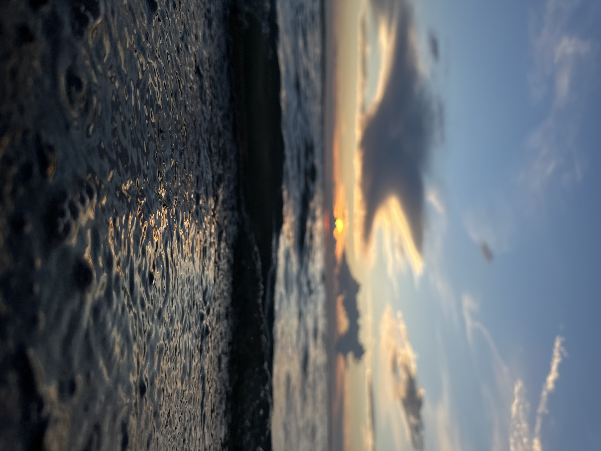 This is a low-angle, close-up shot of shallow water at the edge of a body of water during sunset. The water in the foreground is dark and reflective, showing ripples and small bubbles. In the middle ground, a wave is breaking, creating a darker silhouette against the horizon. The background features the ocean or a large lake with gentle waves and a sunset. The sky is a mix of blue, orange, and pink hues, with scattered clouds. The sun is low on the horizon, partially obscured by a dark cloud formation. The scene evokes a sense of tranquility and natural beauty. The image does not contain any specific landmarks or context to place it in Kokshetau, Kazakhstan.
