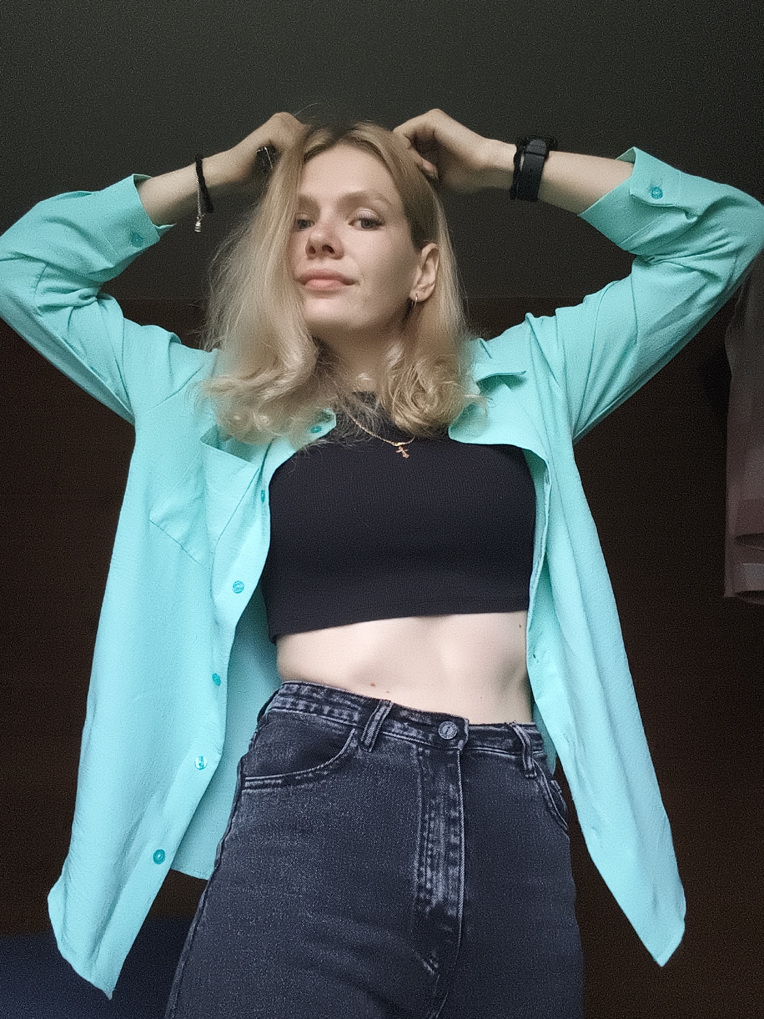 A young woman with blonde hair is posing for a photo, her arms raised above her head and her hands tucked into her hair. She is wearing a teal button-up shirt over a black crop top and dark wash jeans. She has a bracelet on her left wrist and a watch on her right. The background is dark, suggesting an indoor setting. The image does not contain any identifiable landmarks or elements that would indicate a specific location within Русский Вожой, Russia.