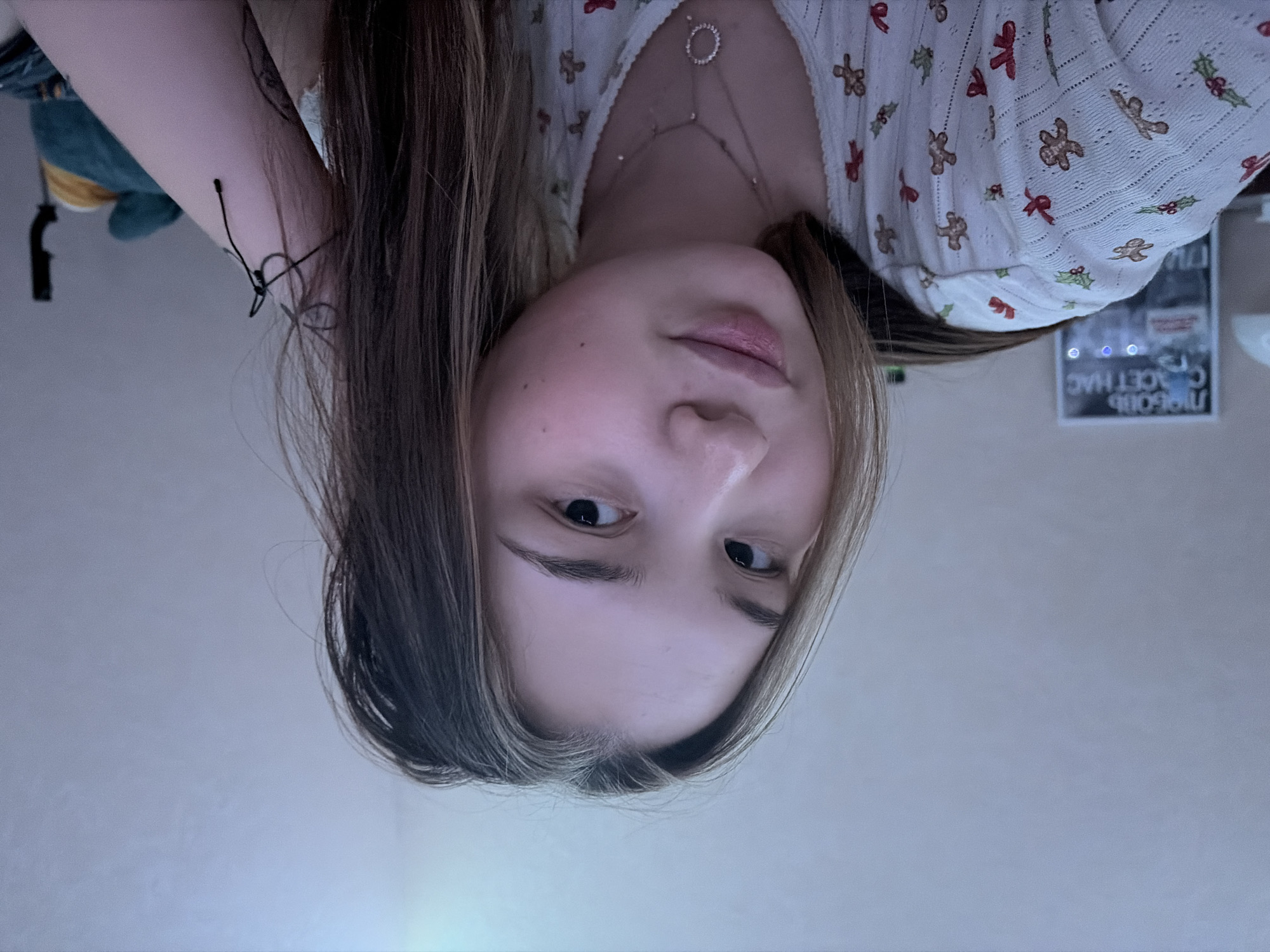 A young woman with long, brown and blonde hair is looking at the camera. She is wearing a white top with a pattern of gingerbread men, holly, and bows. She also wears a necklace. The background shows part of a room with a toy and a sign in Russian. The sign says "ПРИЕХАЛИ В ЧАЙКОВСКИЙ СЪЕДЯТ НАС," meaning "We came to Chaikovsky, they'll eat us."