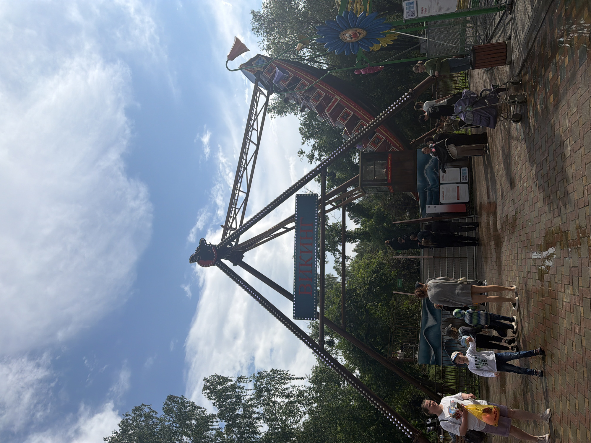 The image depicts an amusement park scene on a partly cloudy day. A large pendulum ride, resembling a pirate ship, is the central attraction, with passengers visible on board. The ride is adorned with lights. A sign with Cyrillic lettering, "ВИКИНГ" (Viking), is prominently displayed on the ride's structure. To the right, a colorful, oversized flower decoration with a face is visible. In the foreground, several people, including adults and children, are gathered on a brick-paved area that shows signs of being wet, possibly from recent rain. Some individuals are standing, while others are walking or attending to a stroller. Trees and foliage surround the amusement park elements, suggesting an outdoor setting. The ground is paved with red and brown bricks, with puddles reflecting the sky.