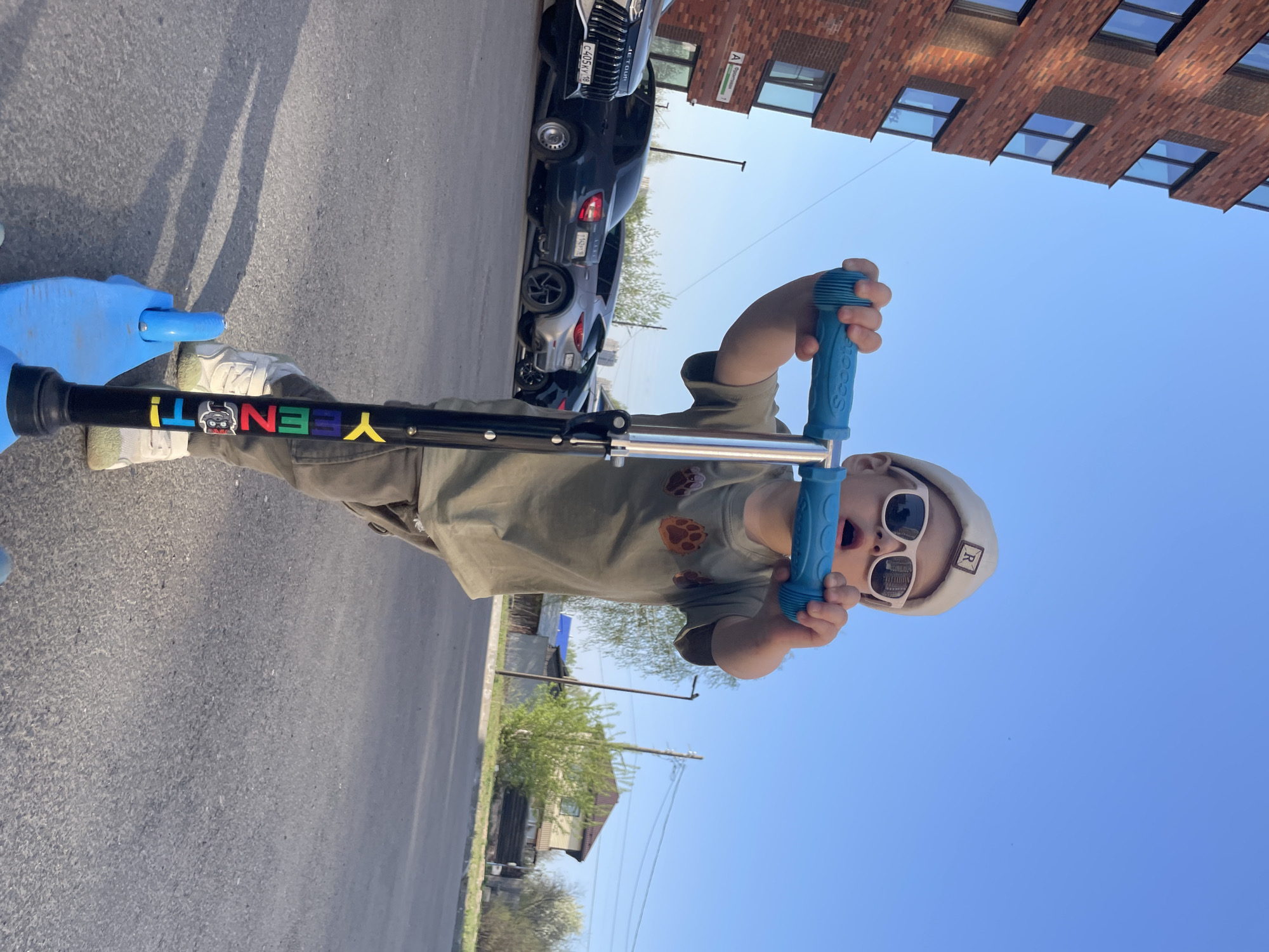 A child wearing sunglasses and a light-colored hat is riding a blue scooter on a paved street. The scooter has "YEENOT!" written on the handlebar stem in multicolored letters. The child is wearing an olive-green shirt with brown paw prints. To the left, cars are parked next to a brick building with multiple windows. In the background, trees and a building with a pointed roof are visible under a blue sky. A street sign marked "A" in Cyrillic characters can be seen on the brick building. Based on these details, the location is identified as Izhevsk, Russia.