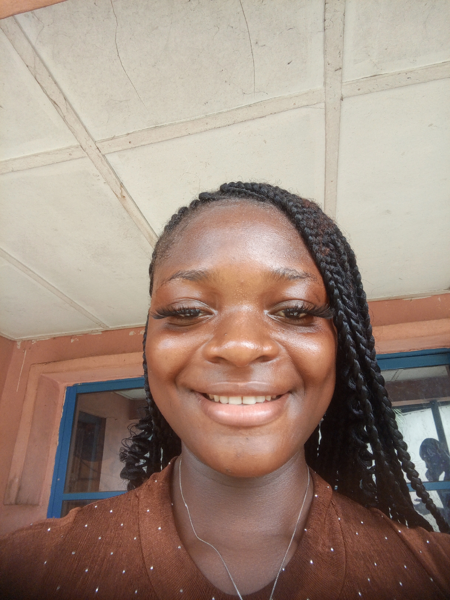 A close-up, self-portrait of a young Black woman with dark braids smiling at the camera. She is wearing a brown top with small, evenly-spaced white dots. A silver necklace is visible around her neck. The background includes an exterior wall with a blue-framed window. Above her is a light-colored, paneled ceiling with thin, wooden crossbeams. The location is indicated by the background and the person's features as being in Uyo, Nigeria.