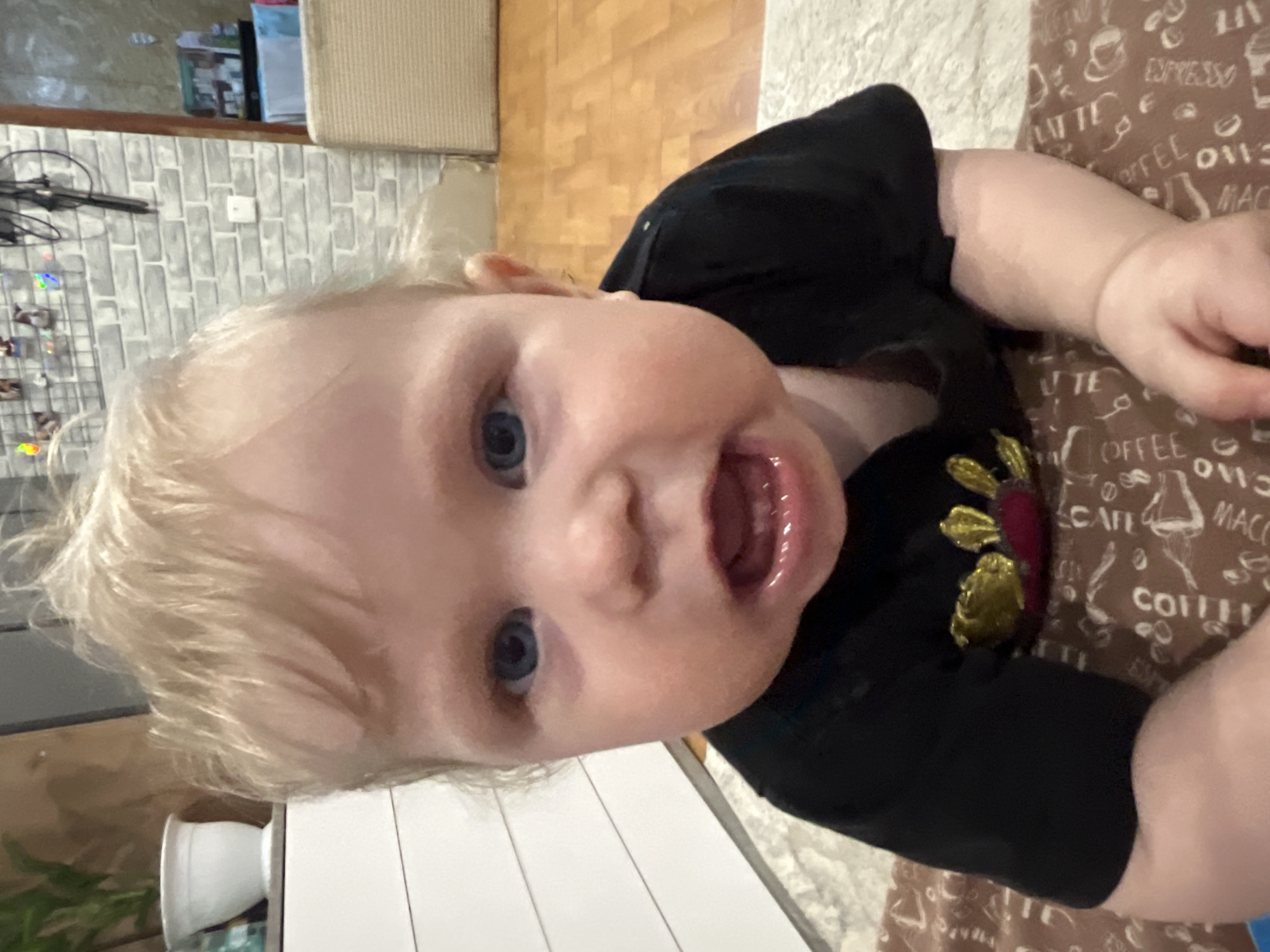 A young child with blonde hair and blue eyes is lying on a surface covered with a brown patterned fabric. The child is wearing a black garment with a gold flower embellishment. The background includes a wooden floor, a brick wall, and a white cabinet or countertop. The location is not determinable.